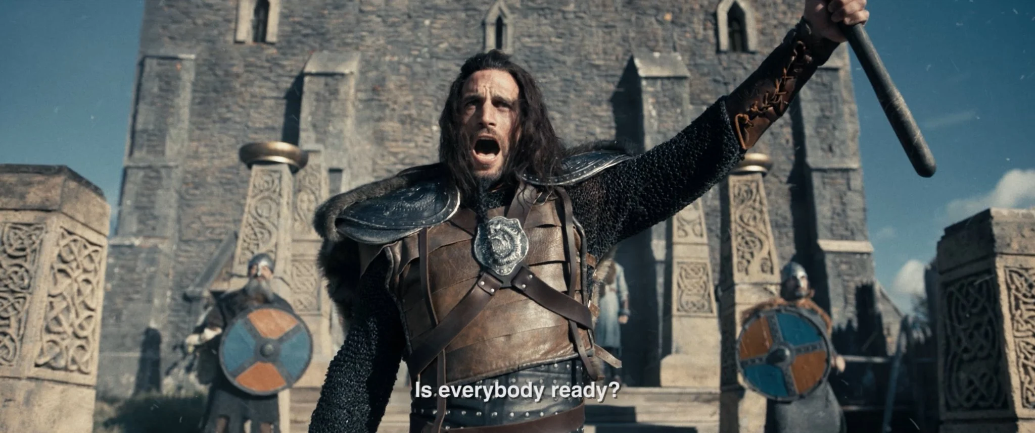 A man dressed as a medieval warrior, wearing armor and chainmail, raising his sword, standing in front of a stone castle with guards holding shields on either side. Subtitles read, 'Is everybody ready?'