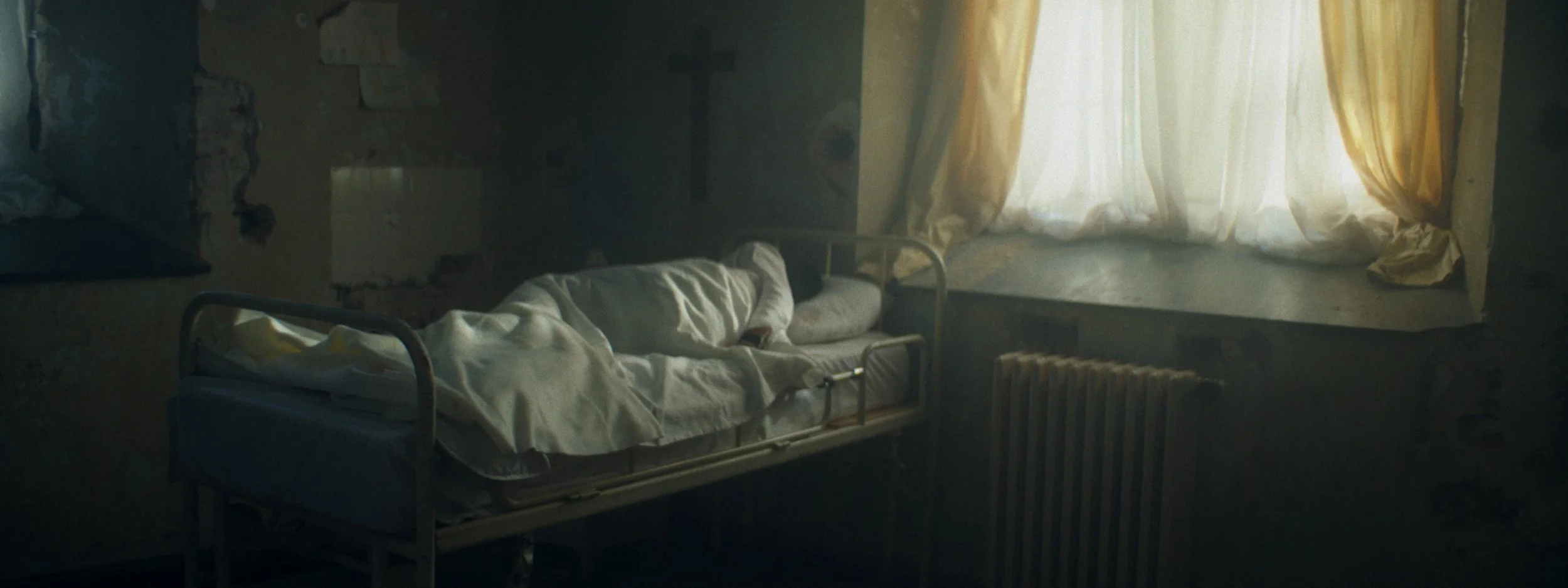 A person lying on a hospital bed in a dimly lit room with yellow curtains and a radiator.