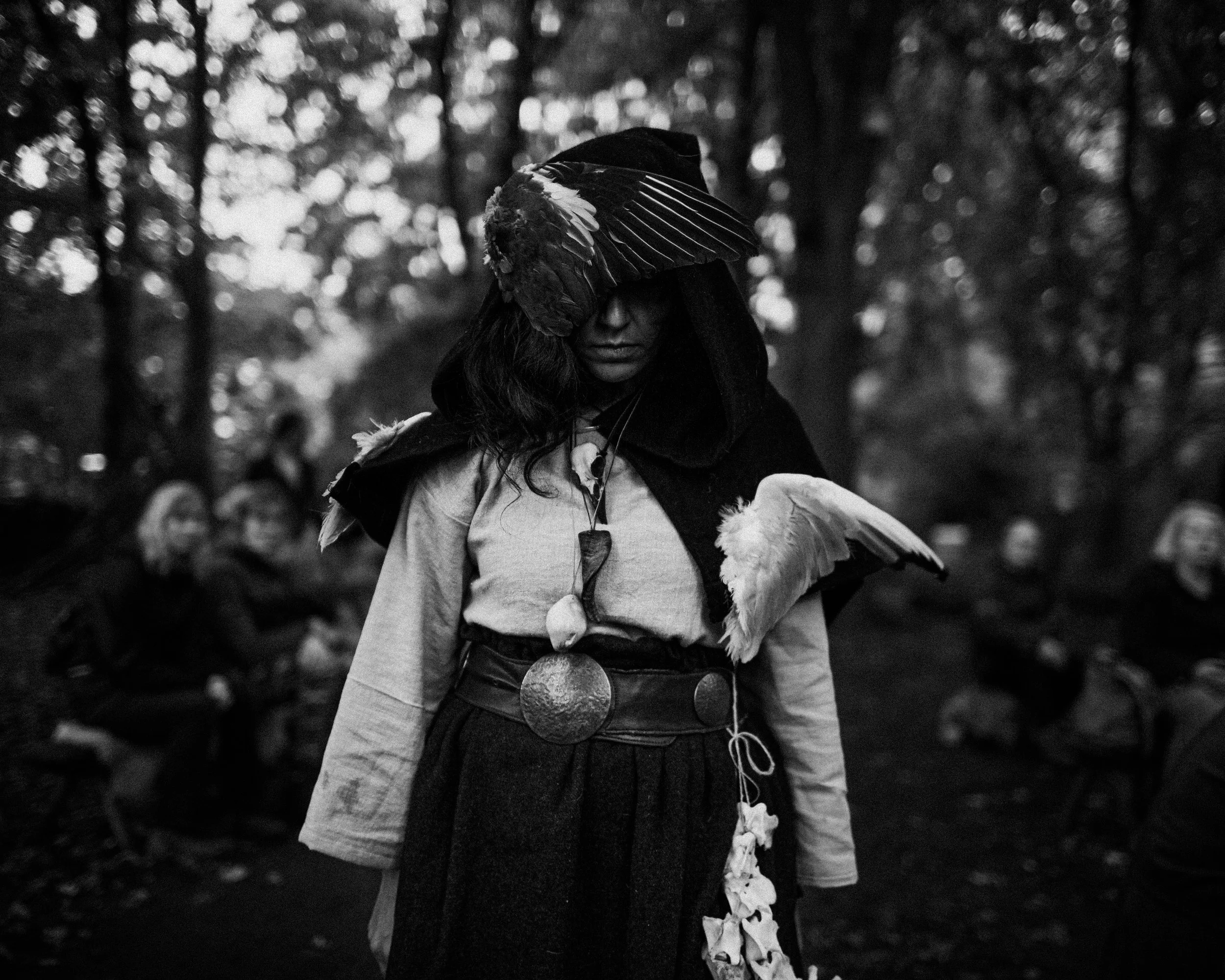 A woman in dark clothing with long hair wears a large feathered headdress and stands in a forest, with a group of children in the background.