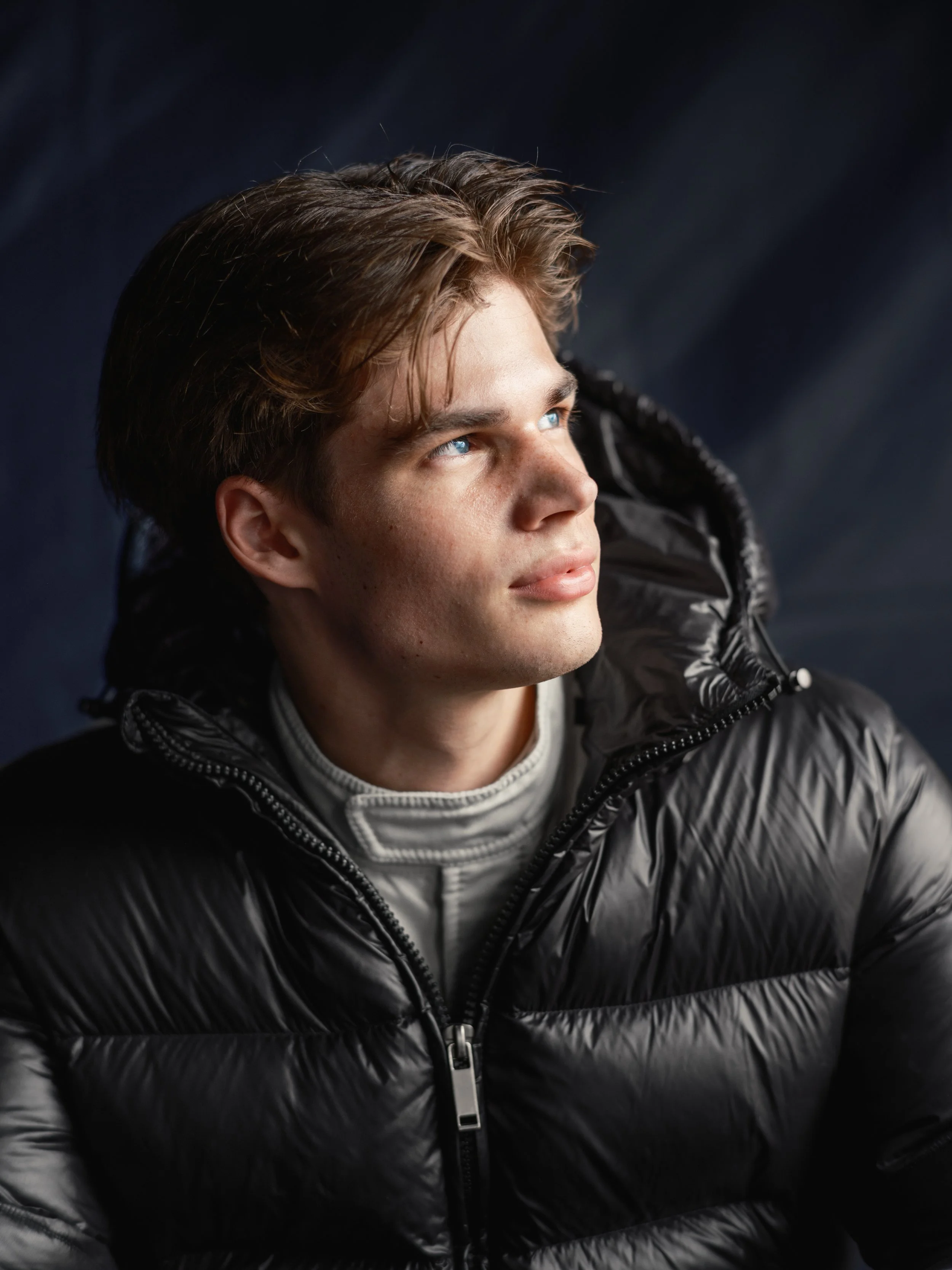 A young man with light skin, blue eyes, and light brown hair, wearing a black puffy jacket, looking to the right against a dark background.