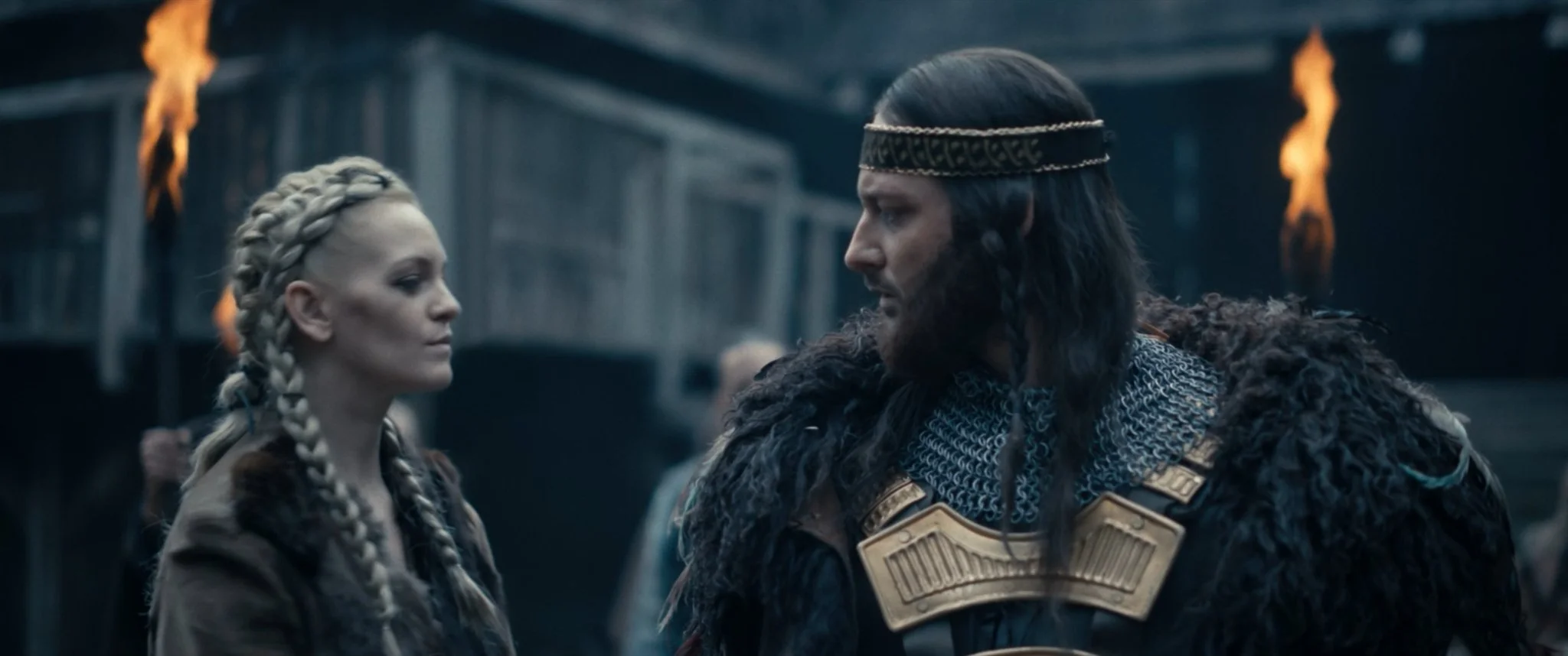 Two characters dressed in medieval or fantasy costumes facing each other, with a background of flames and a dark structure. One is a woman with braided blonde hair, and the other is a man with long hair, a beard, and a headband, both wearing armor.