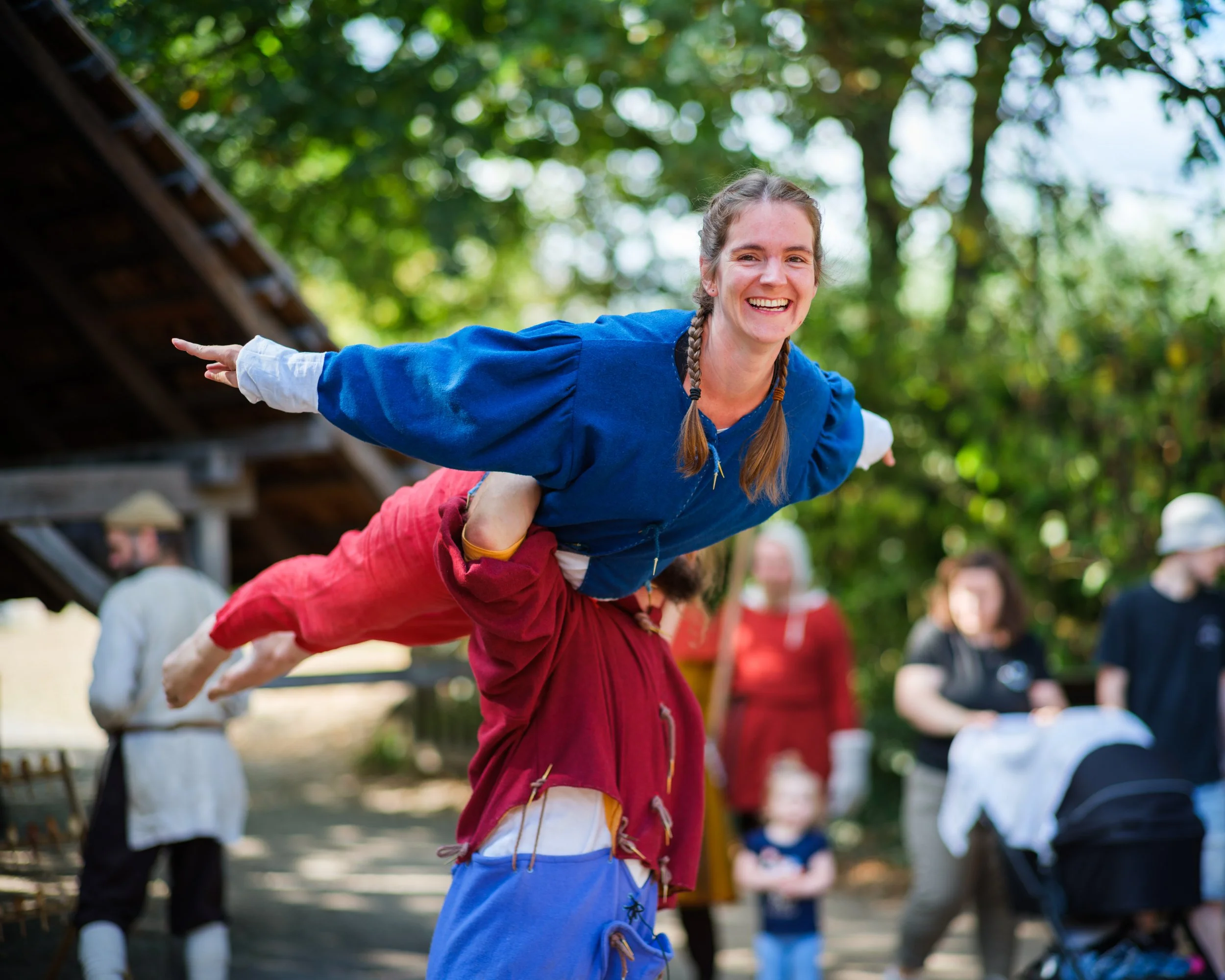 A woman with a braid and blue shirt balancing on a person's back during an outdoor event, smiling with trees and people in the background.