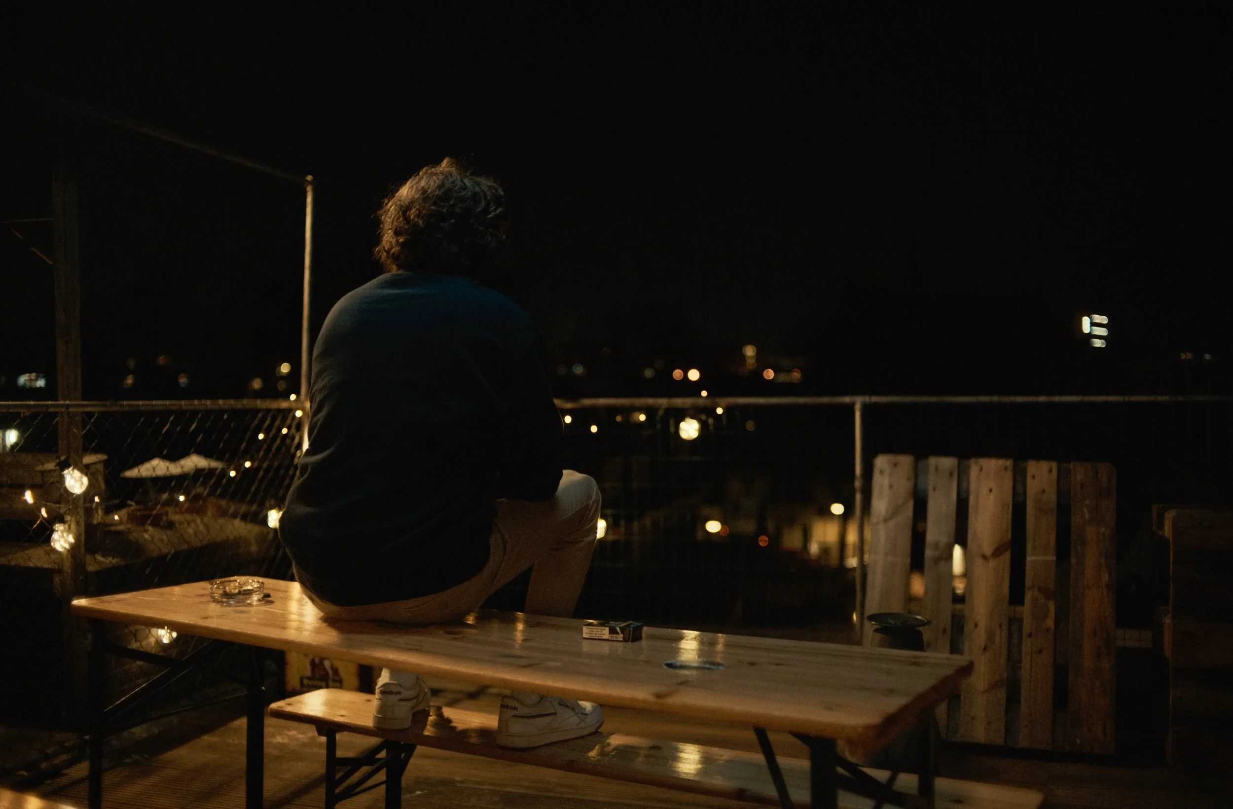 Person sitting on a wooden table at night, facing away, with city lights and a dark sky in the background, smoking or holding something in hand.