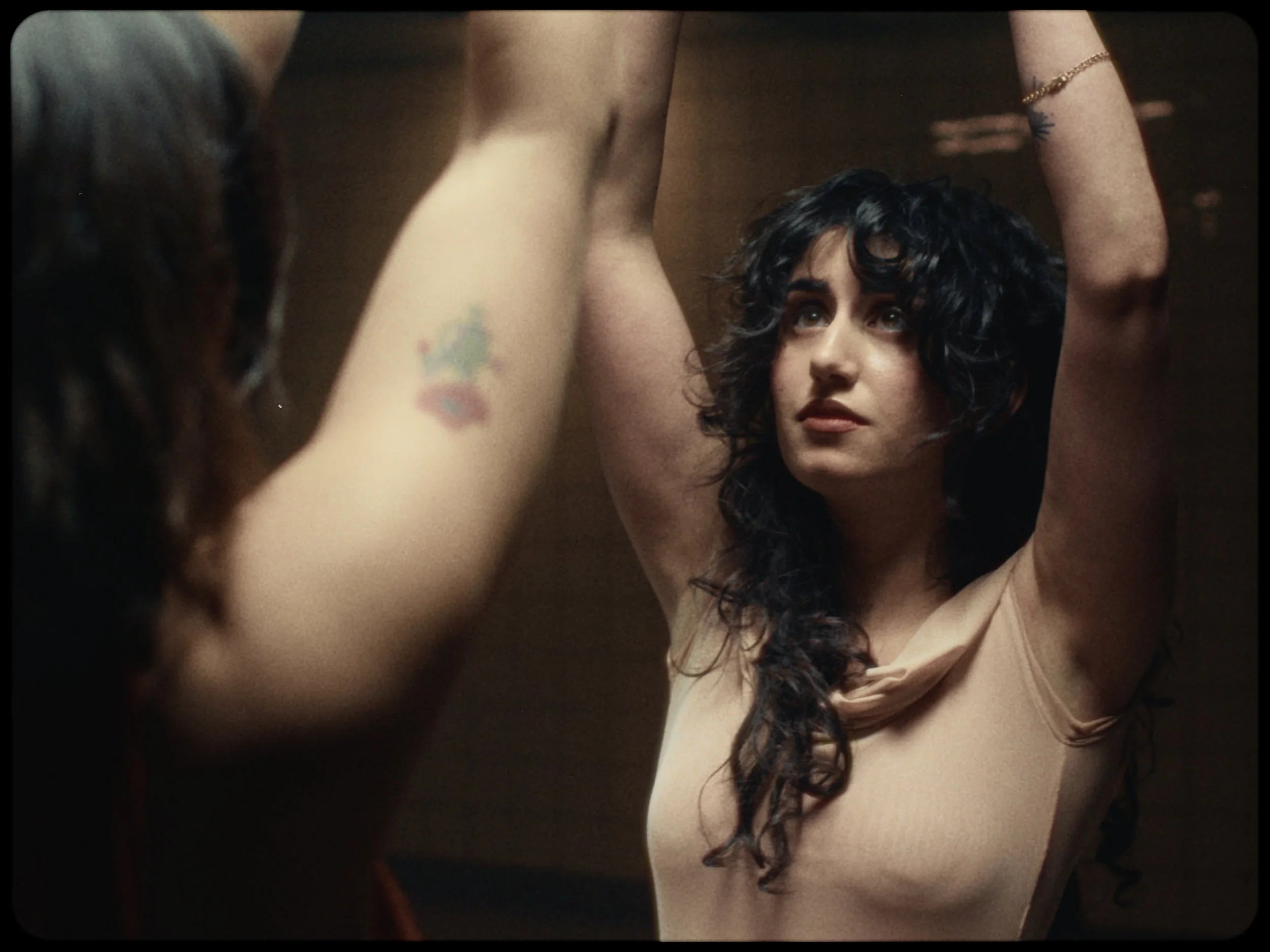 A woman with curly dark hair and tattoos on her arm looks up while posing with her arms raised, revealing her underarms and a beige top.