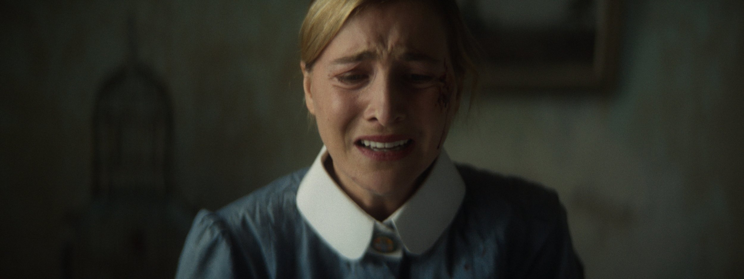 A woman with blonde hair, wearing a dark shirt with a white collar, looking distressed with a pained expression.
