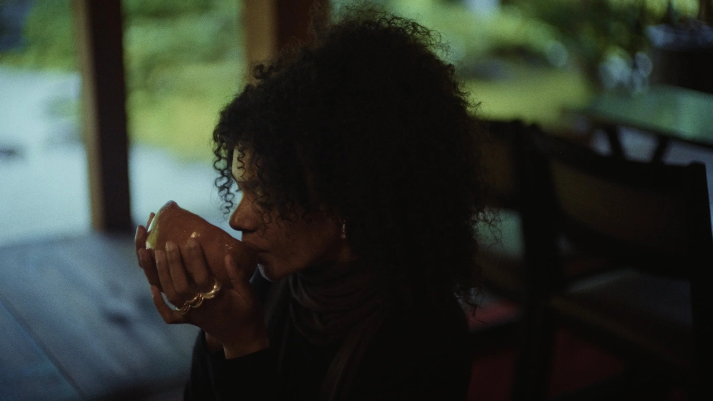 A woman with curly hair drinking from a bowl or cup, sitting in a dimly lit room with a large window showing greenery outside.