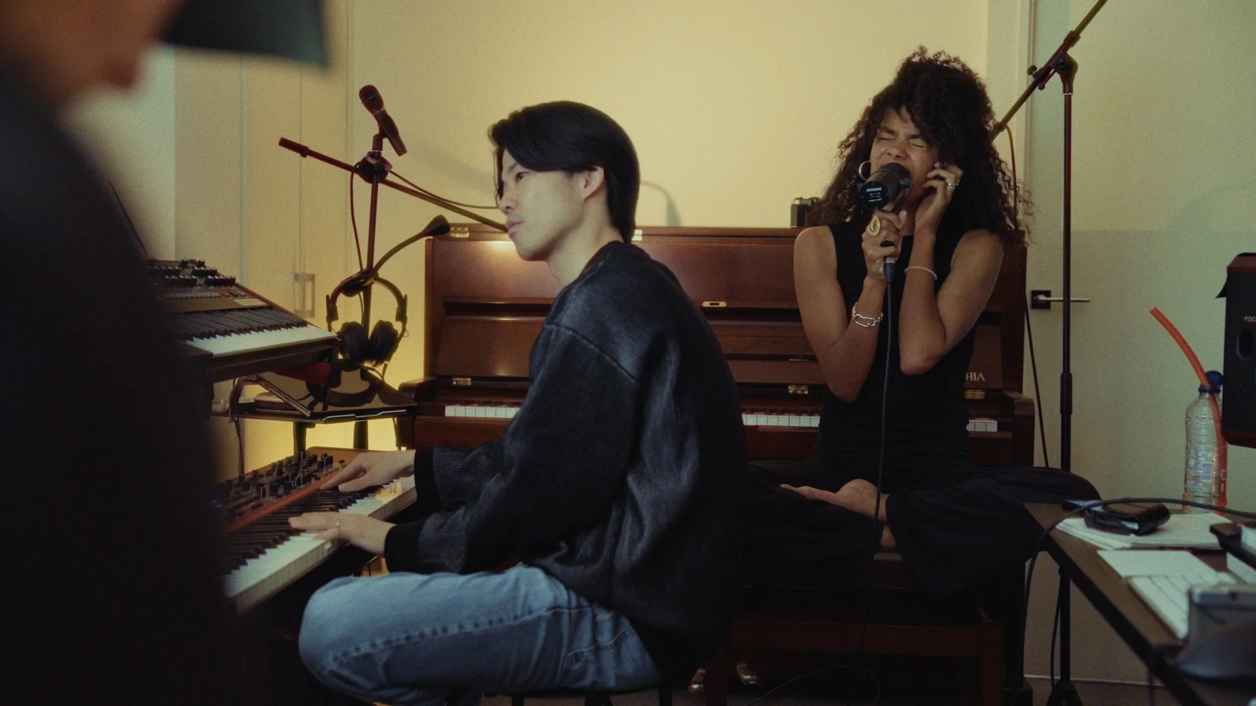 A woman singing into a microphone while a man plays a keyboard in a music studio.