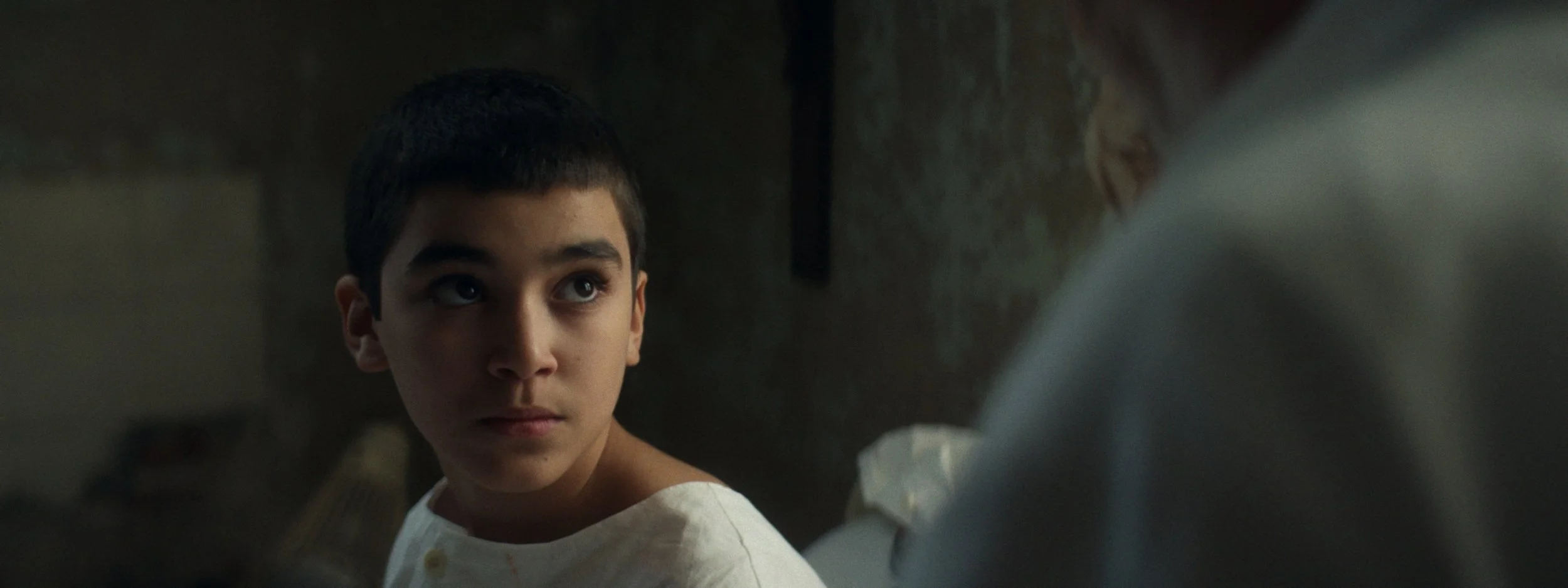 A young boy with short dark hair looking up with a serious expression, sitting indoors with a blurred background.