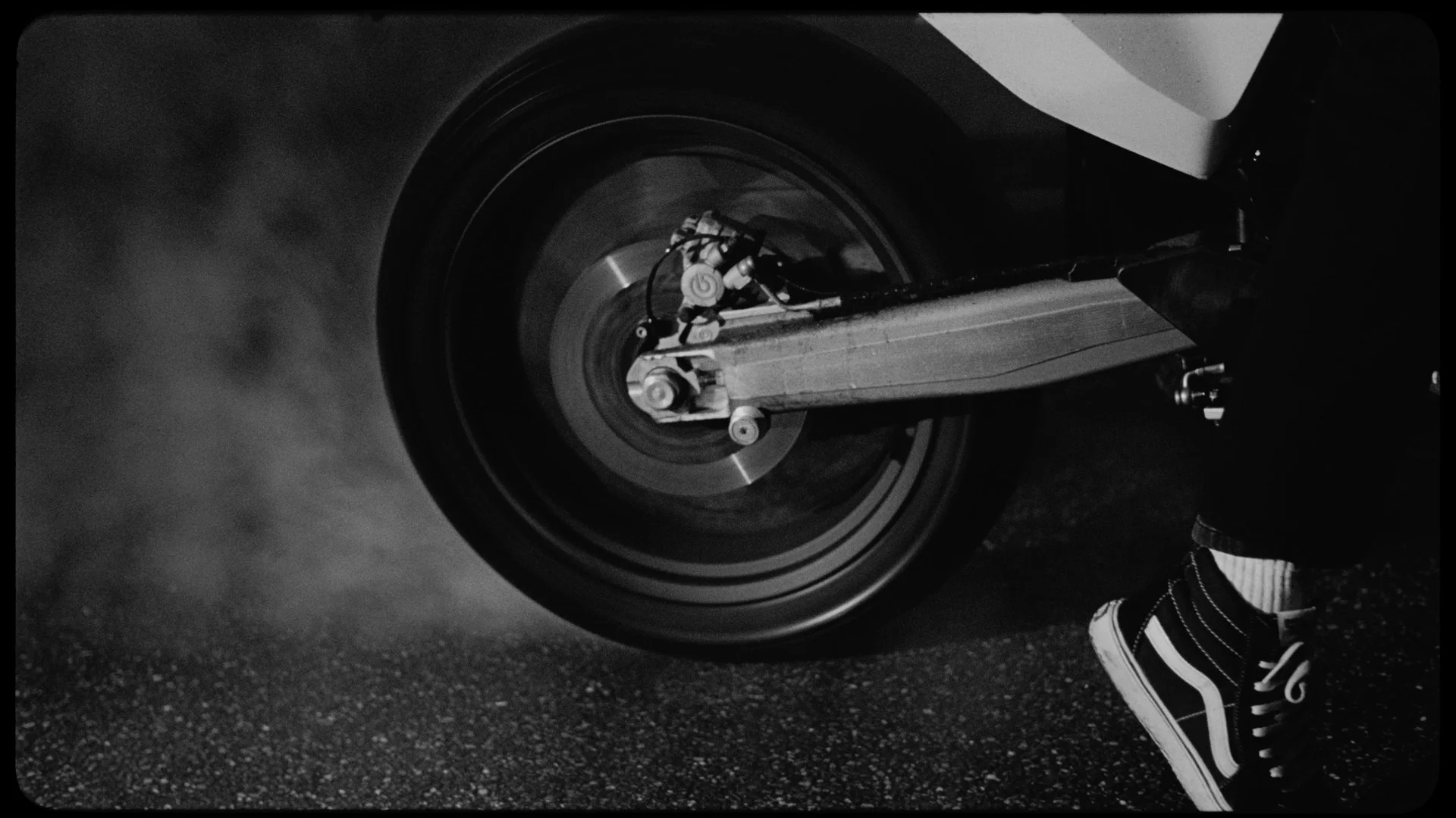 Black and white photo of a car tire in motion next to a person's foot wearing a sneaker.