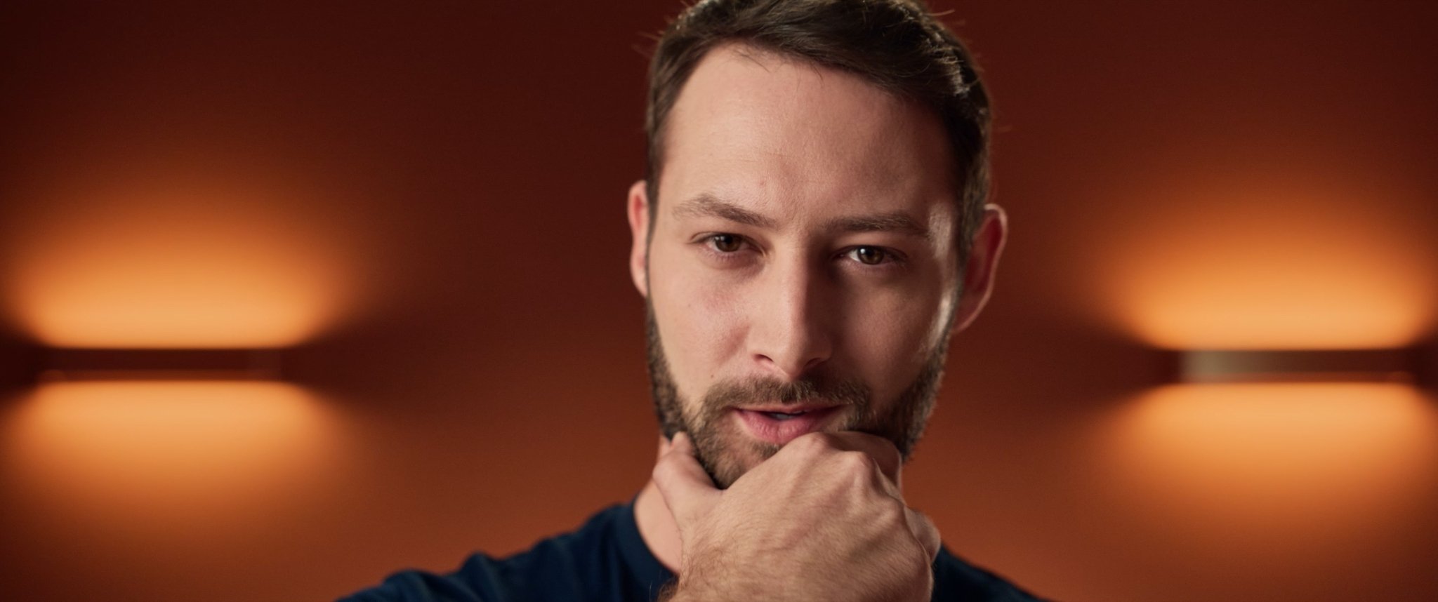 Close-up of a man with a beard looking at the camera with a thoughtful expression, hand near chin, with warm, blurred lights in the background.