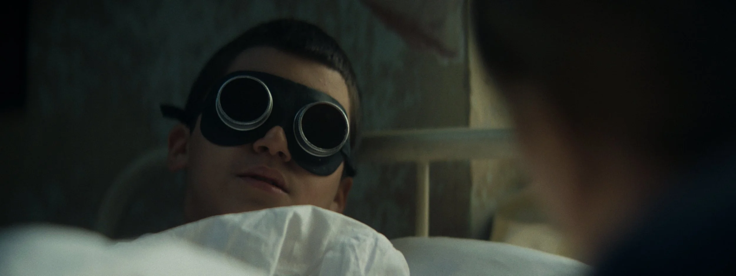 A young boy lying on a bed wearing dark goggles, looking at someone out of the frame with a slight smile.
