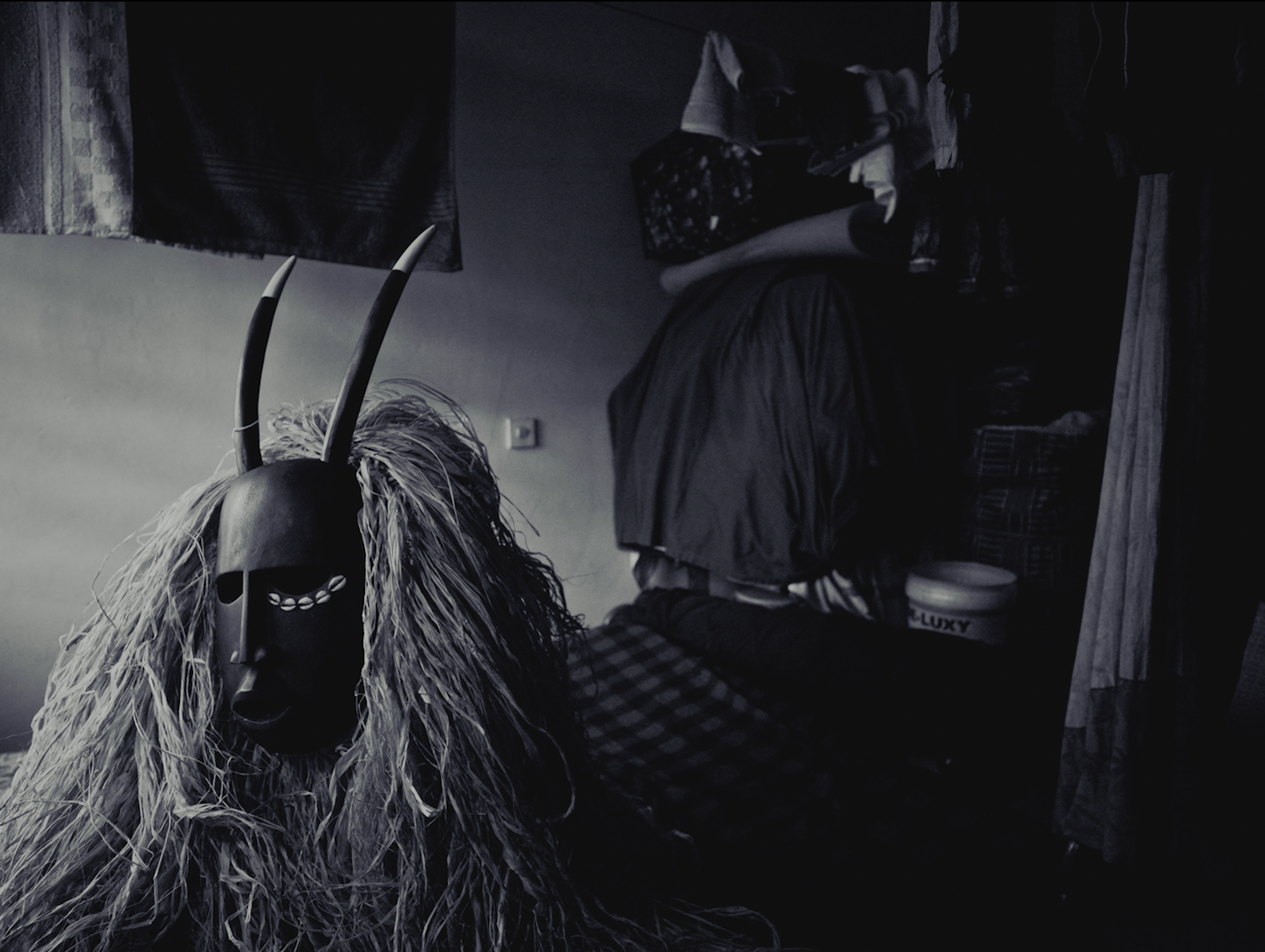 A dark room with a cluttered desk or table, featuring a children’s or plush toy with rabbit ears, surrounded by various fabric items, a pair of legs in striped pants, and some patterned clothing or blankets.