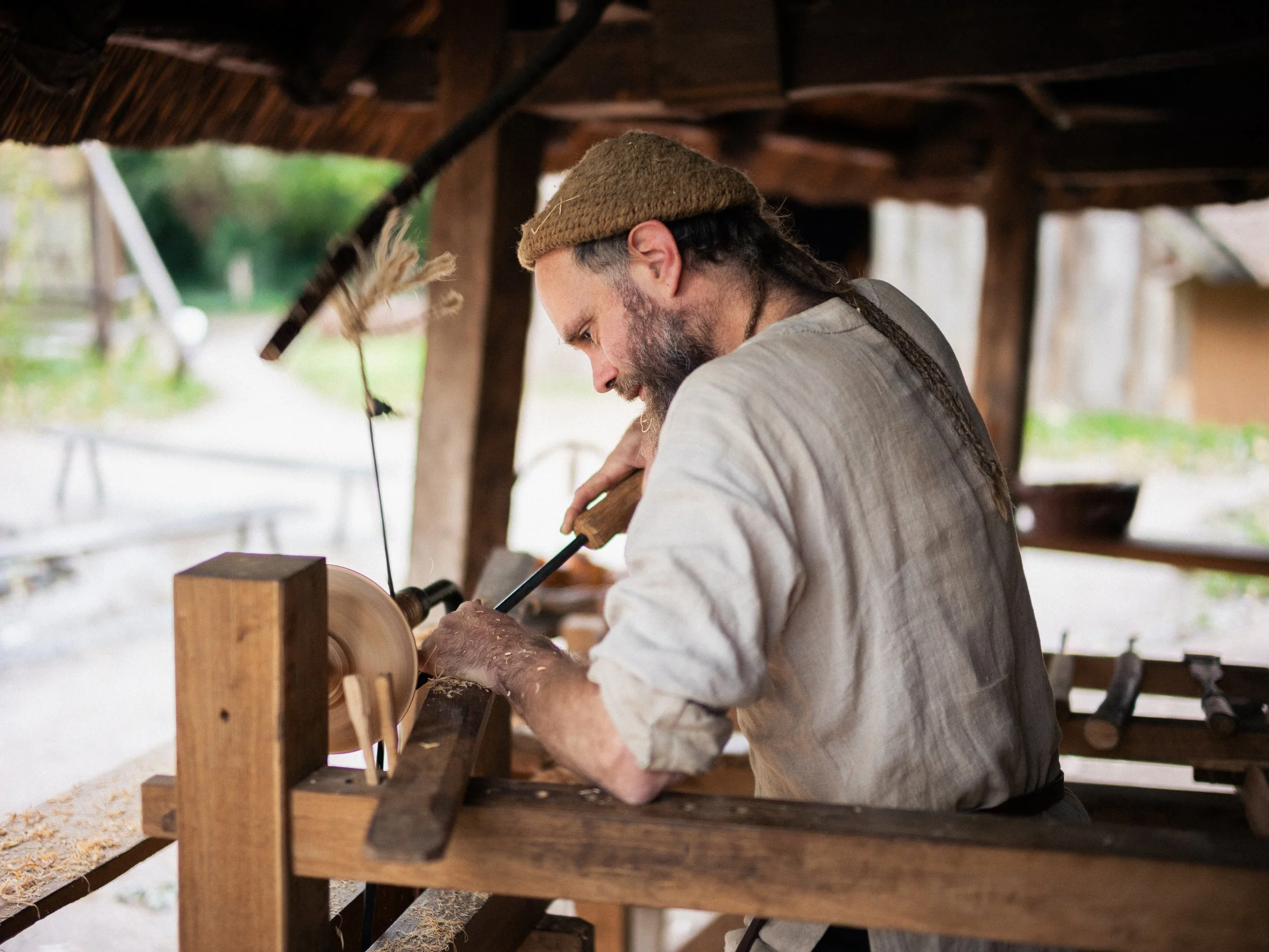 A man with long dreadlocks, wearing a brown knit cap and beige shirt, working on a piece of wood in a rustic workshop with a thatched roof.