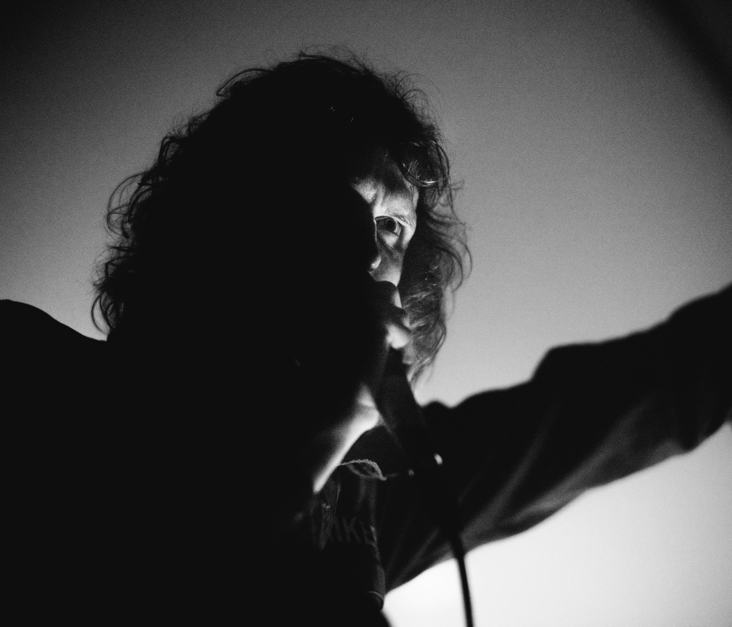 A black-and-white photo of a person with curly hair, looking at the camera, with a shadowed face and a plain background.