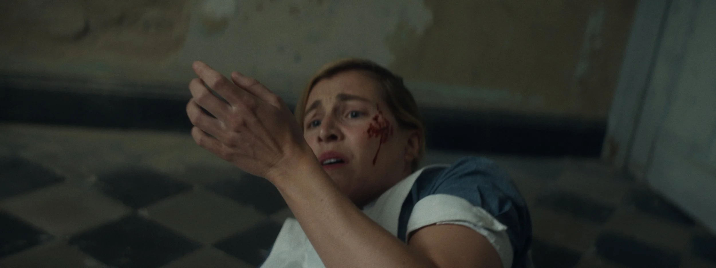 A woman with a bloody face and hands looks distressed as she holds her hands near her face in a checkered floor room.