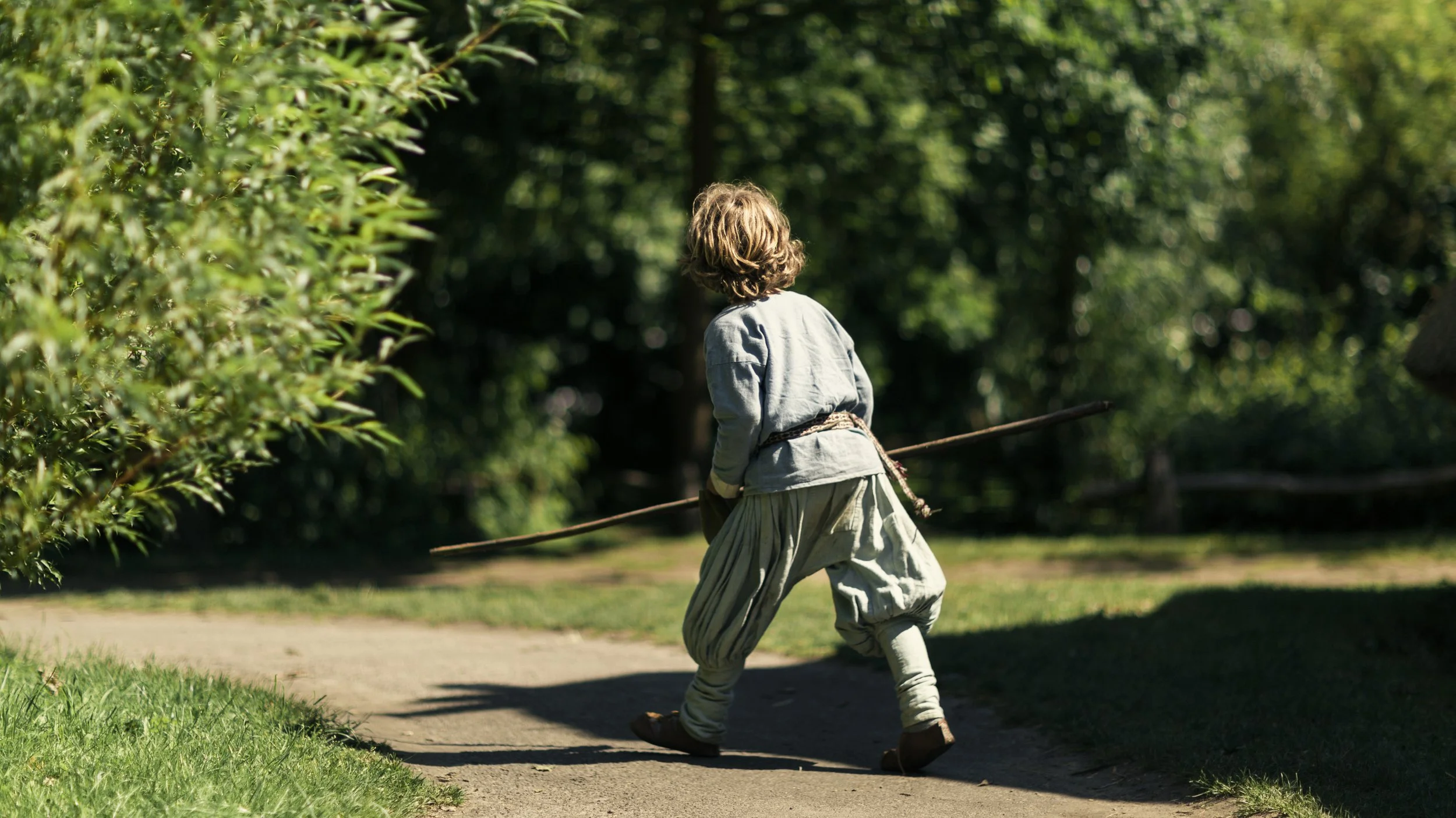 A child dressed in historical or medieval clothing, holding a stick, walking along a dirt path in a wooded park area.
