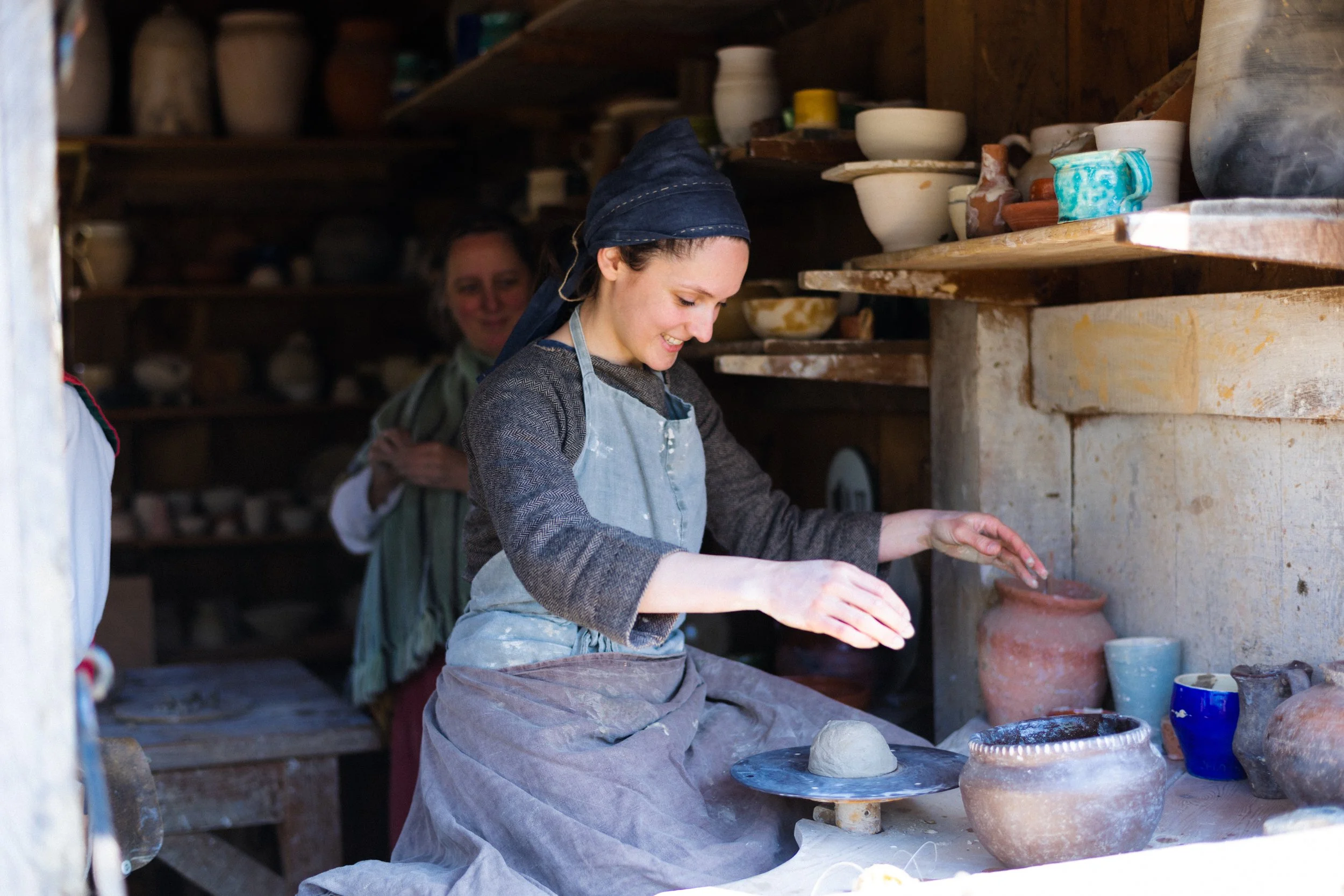 A woman wearing a headscarf and apron working on pottery at a pottery studio. She is shaping clay with her hands, surrounded by various finished and unfinished pottery pieces on shelves around her.