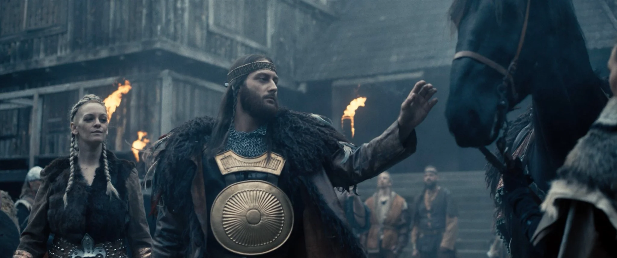 Scene with medieval characters wearing armor and fur, engaging in a ritual or ceremony, with torches burning in the background.