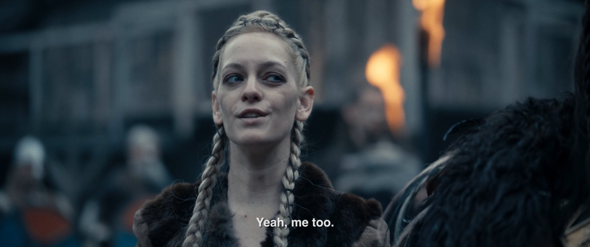 A young woman with long blonde braided hair wearing a fur-lined coat, standing outdoors in a medieval setting with armored soldiers in the background, speaking with someone off-camera.