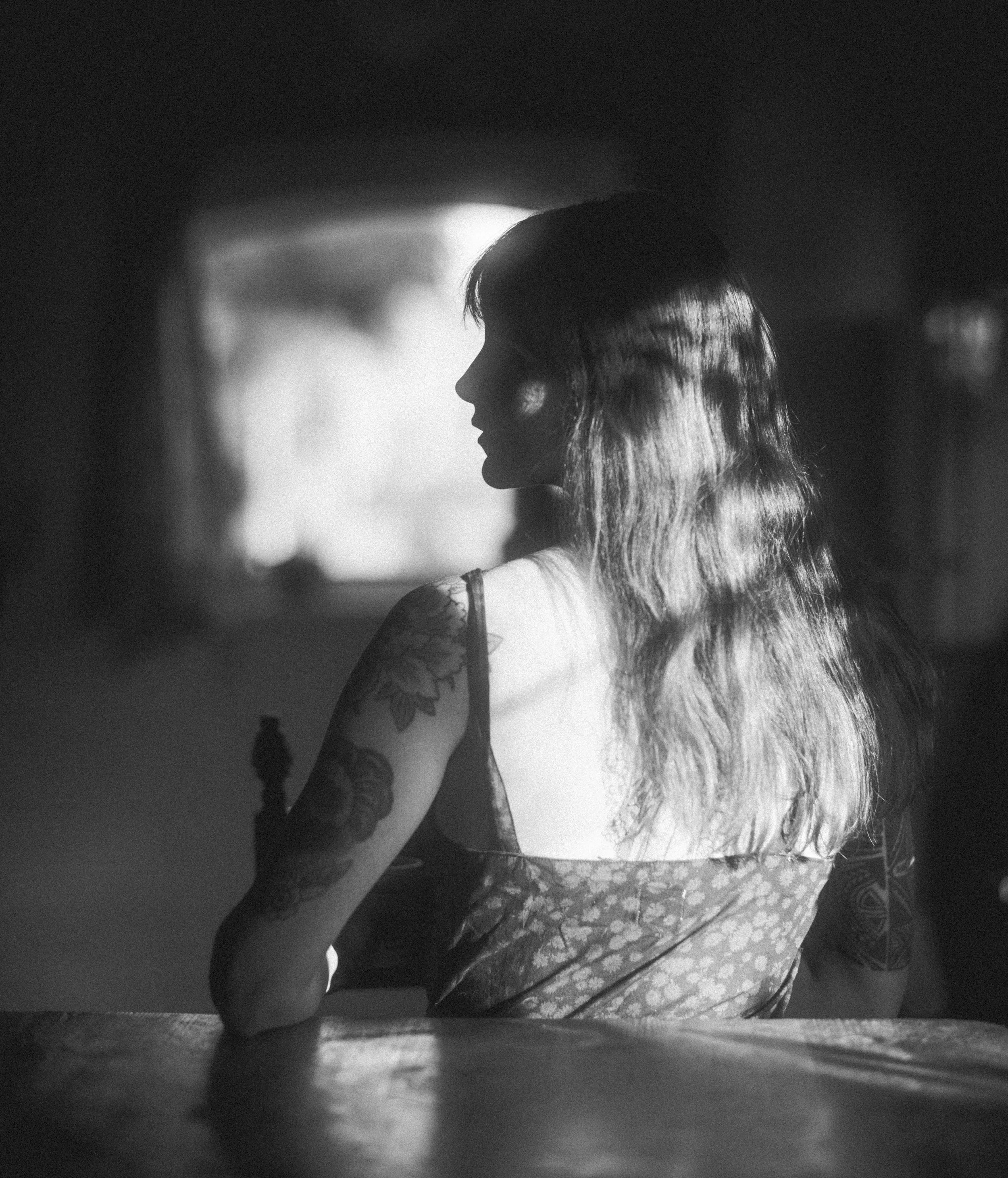 Black and white photo of a woman with long hair, wearing a lace top, posing with her hand under her chin, looking away from the camera.