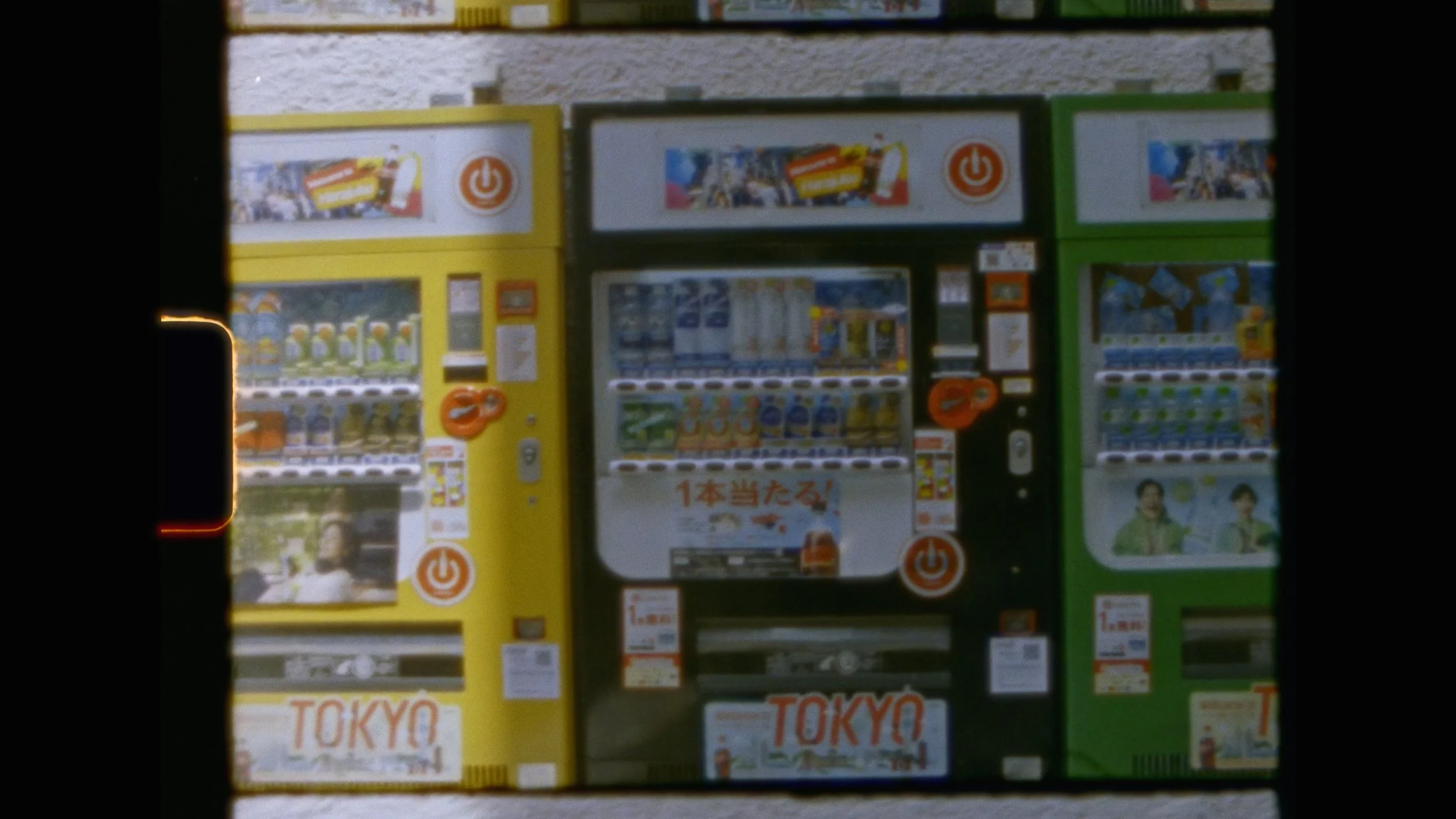 Three vending machines in yellow, black, and green colors, with various snacks and beverages, including some with colorful packaging, and Japanese text on some of the machines.