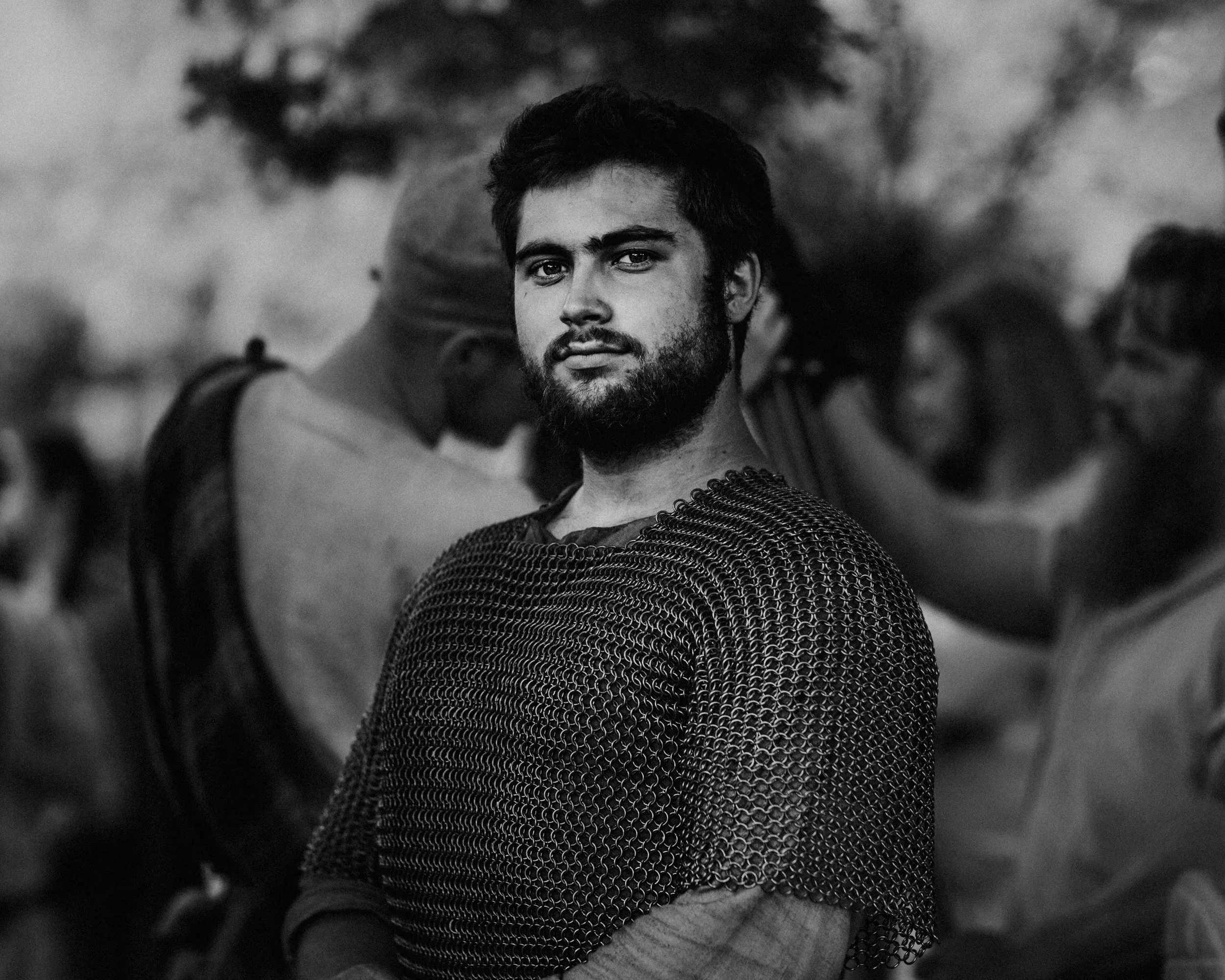 A young man with a beard and short hair looking into the camera, standing outdoors with trees in the background. The photo is in black and white.