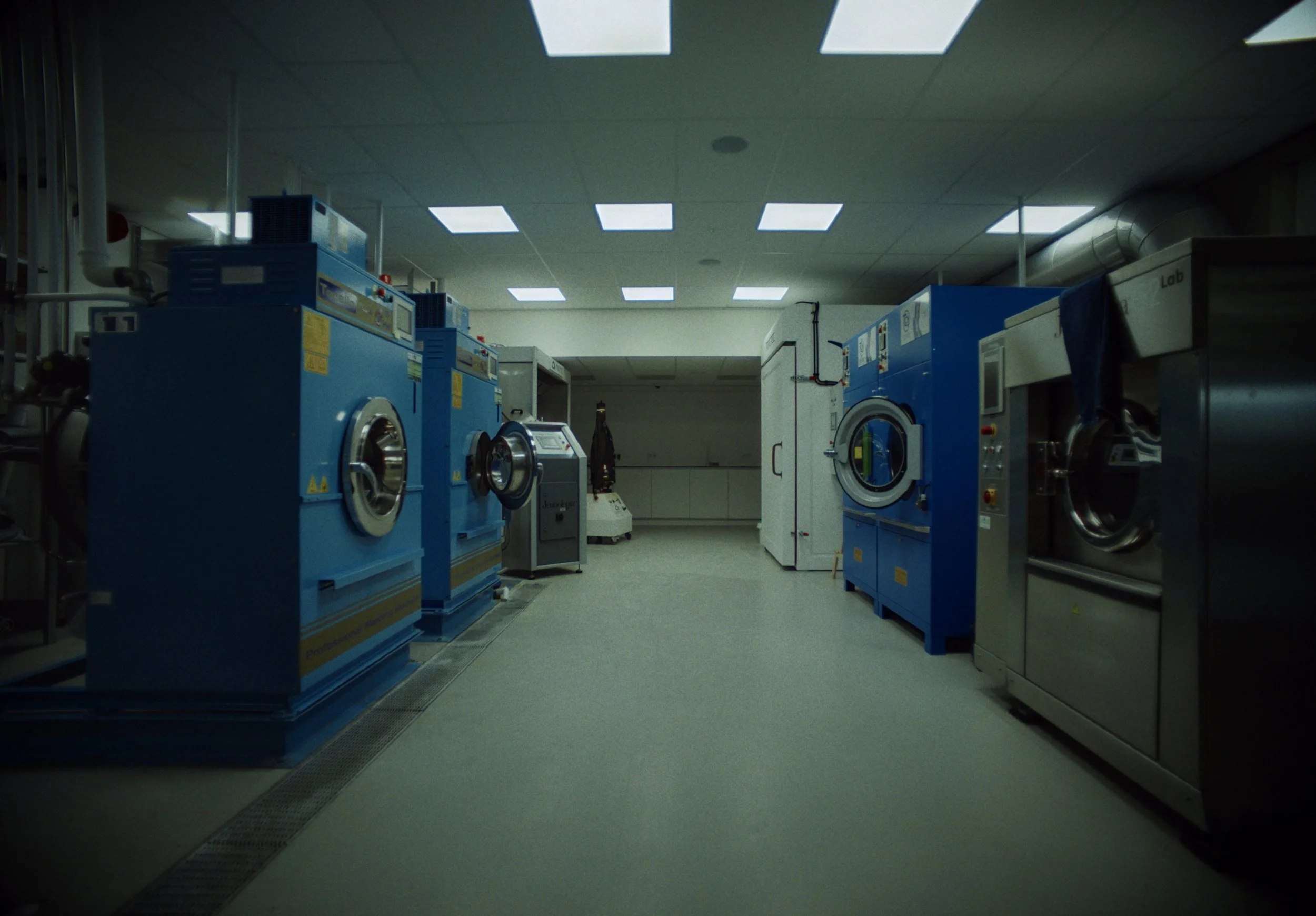 Laundry room with blue commercial washing machines and dryers, fluorescent ceiling lights, and a laundry cart in the background.