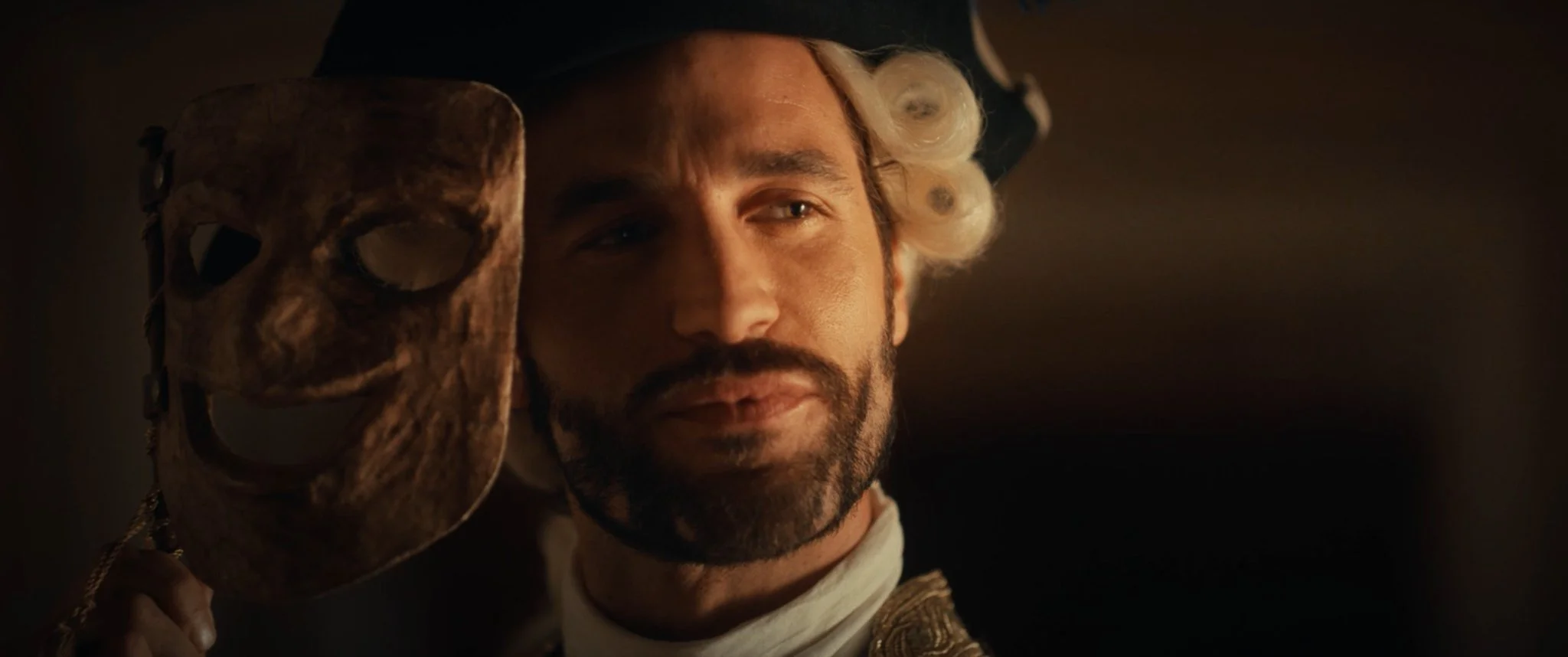 A man in historical costume holding a wooden mask next to his face.