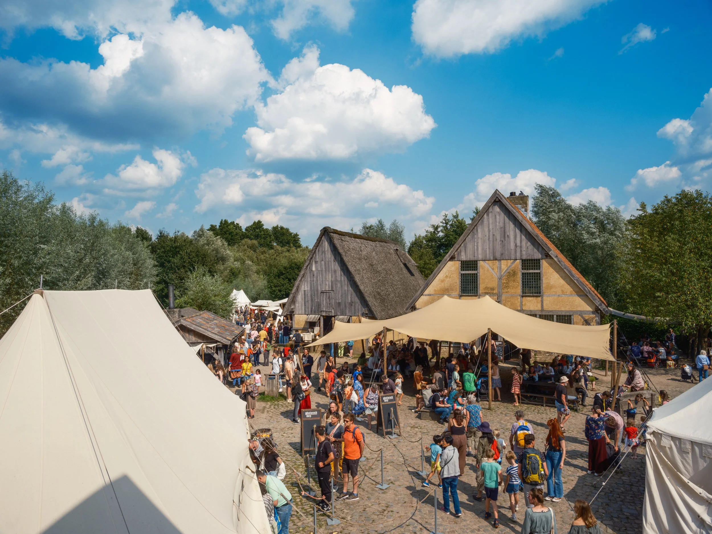 People gathering at an outdoor event with tents and rustic buildings under a cloudy sky.