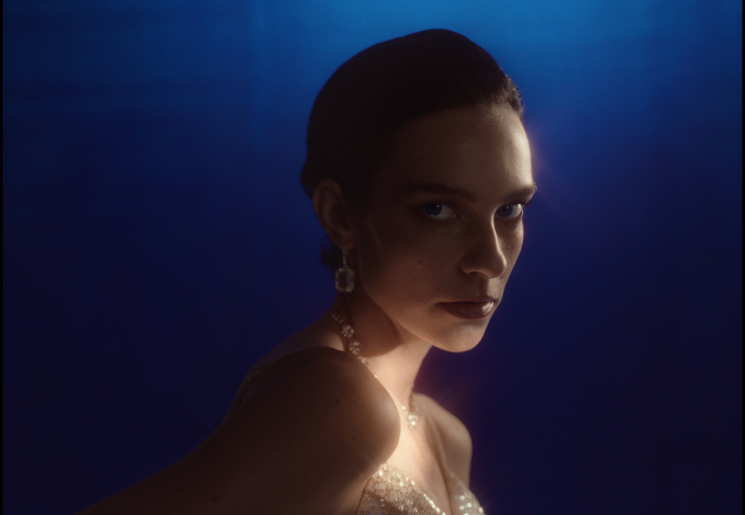 A woman with dark hair styled back, wearing pearl earrings and a necklace, is dressed in a sparkly dress, with a blue gradient background.