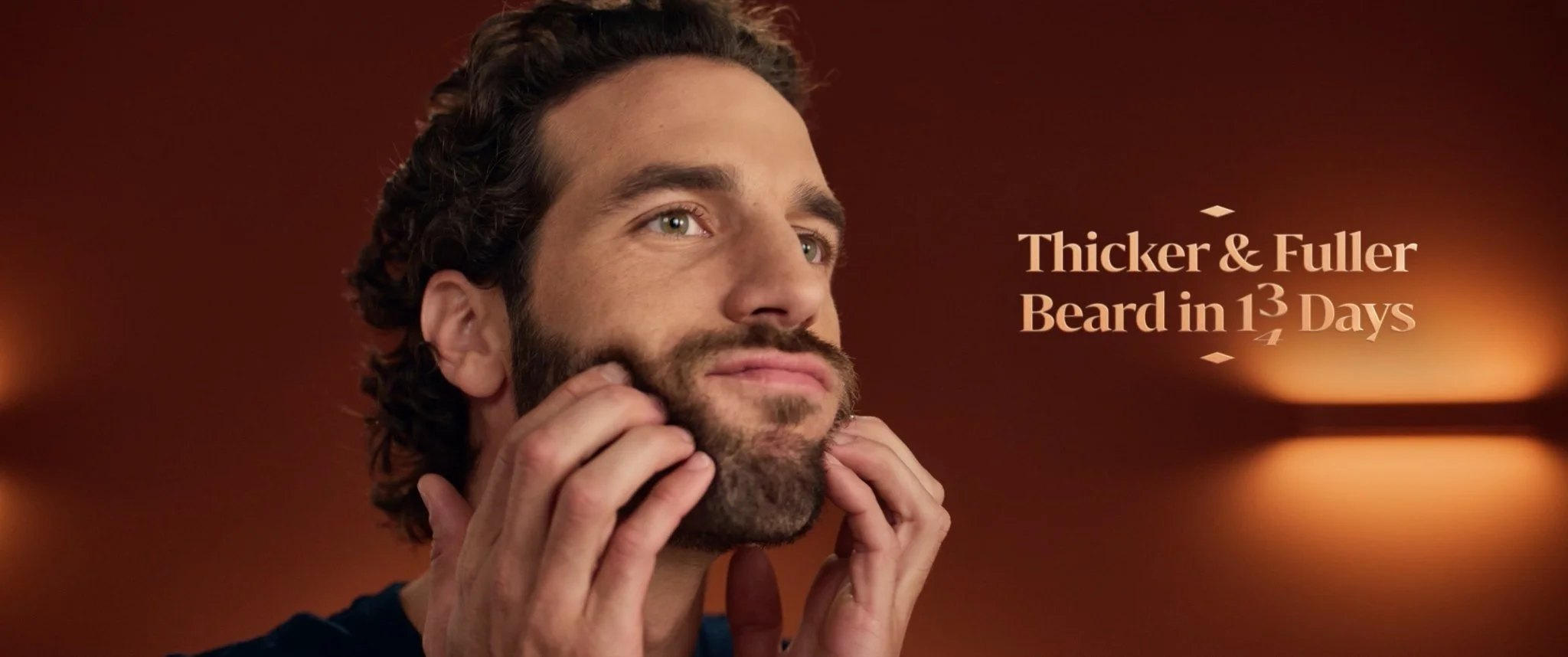 A man with dark hair and a beard is touching his beard with two hands. The background is warm-colored with blurred lights, and there is text that reads 'Thicker & Fuller Beard in 13 Days'.