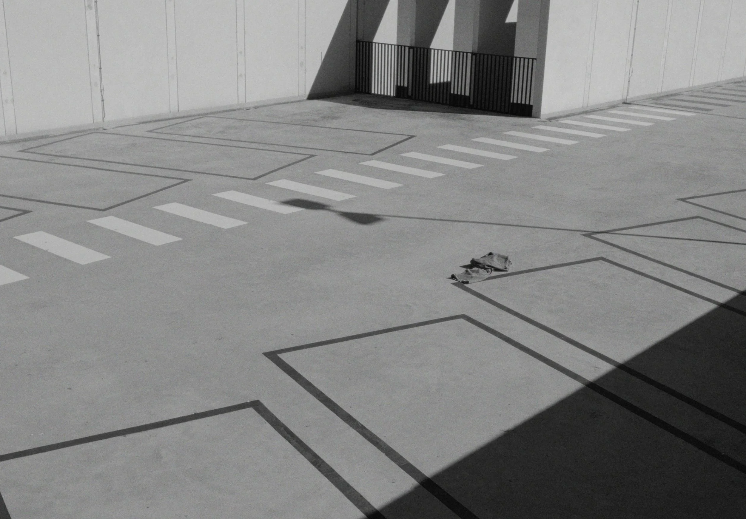Empty parking lot with marked parking spaces, a small trash bag on the ground, and a wall with shadows cast by a fence.