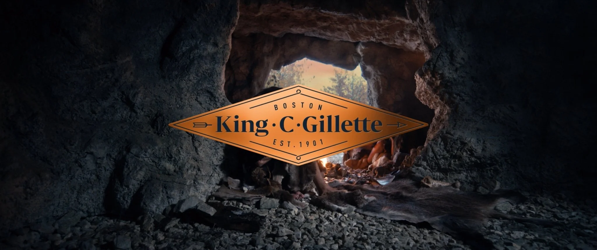 A view from inside a rock cave looking outwards at trees and sky, with an orange sign in the foreground reading "Boston King C. Gillette Est. 1901."