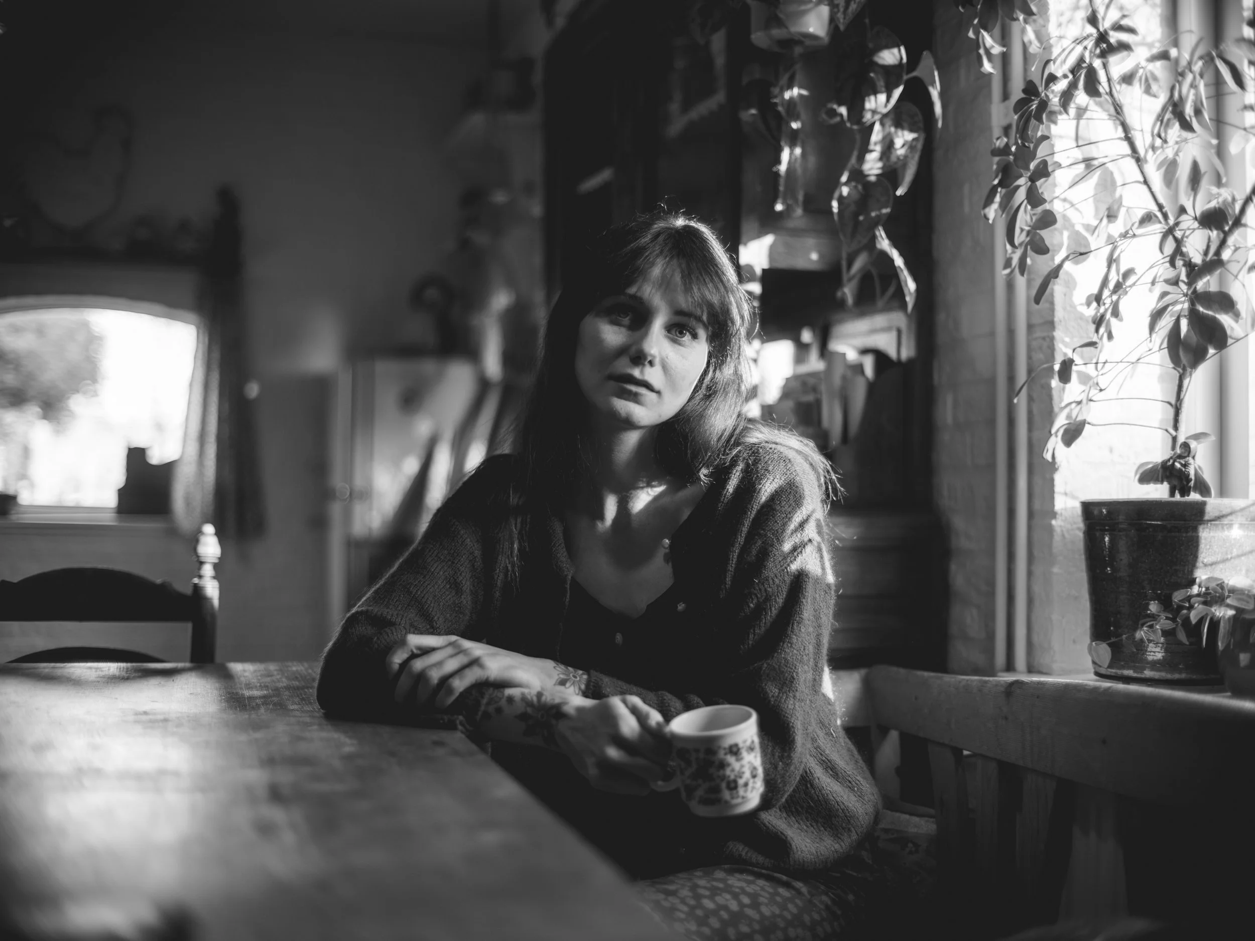 A woman with long dark hair and tattoos on her arms sitting at a wooden table, holding a coffee mug, indoors with plants and sunlight streaming through a window.