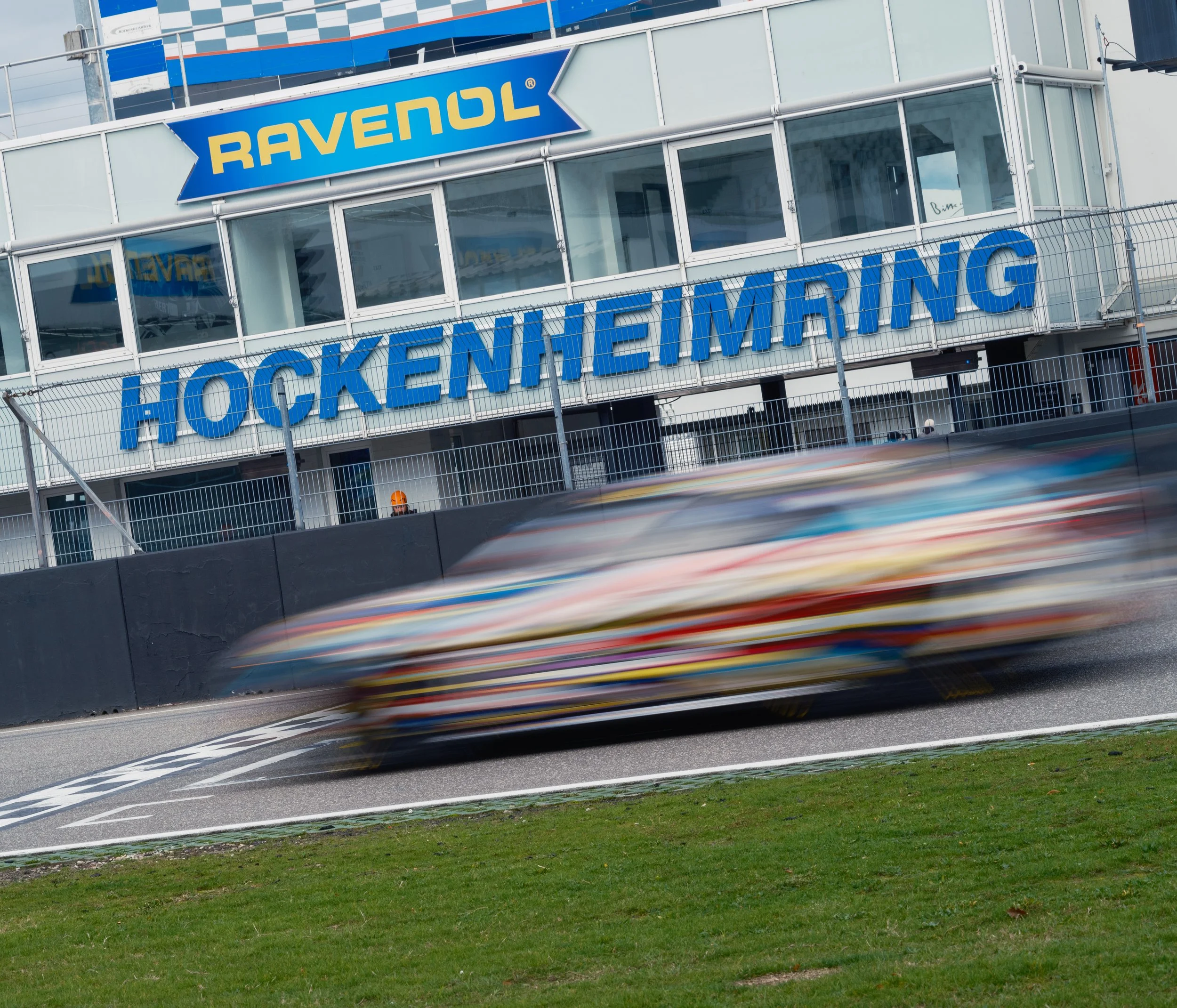 A blurred race car speeding on a racetrack in front of a modern glass building with large blue and yellow lettering that reads 'RAVENOL' and 'HOCKENHEIM RING.' A person wearing an orange helmet is visible behind the fence.