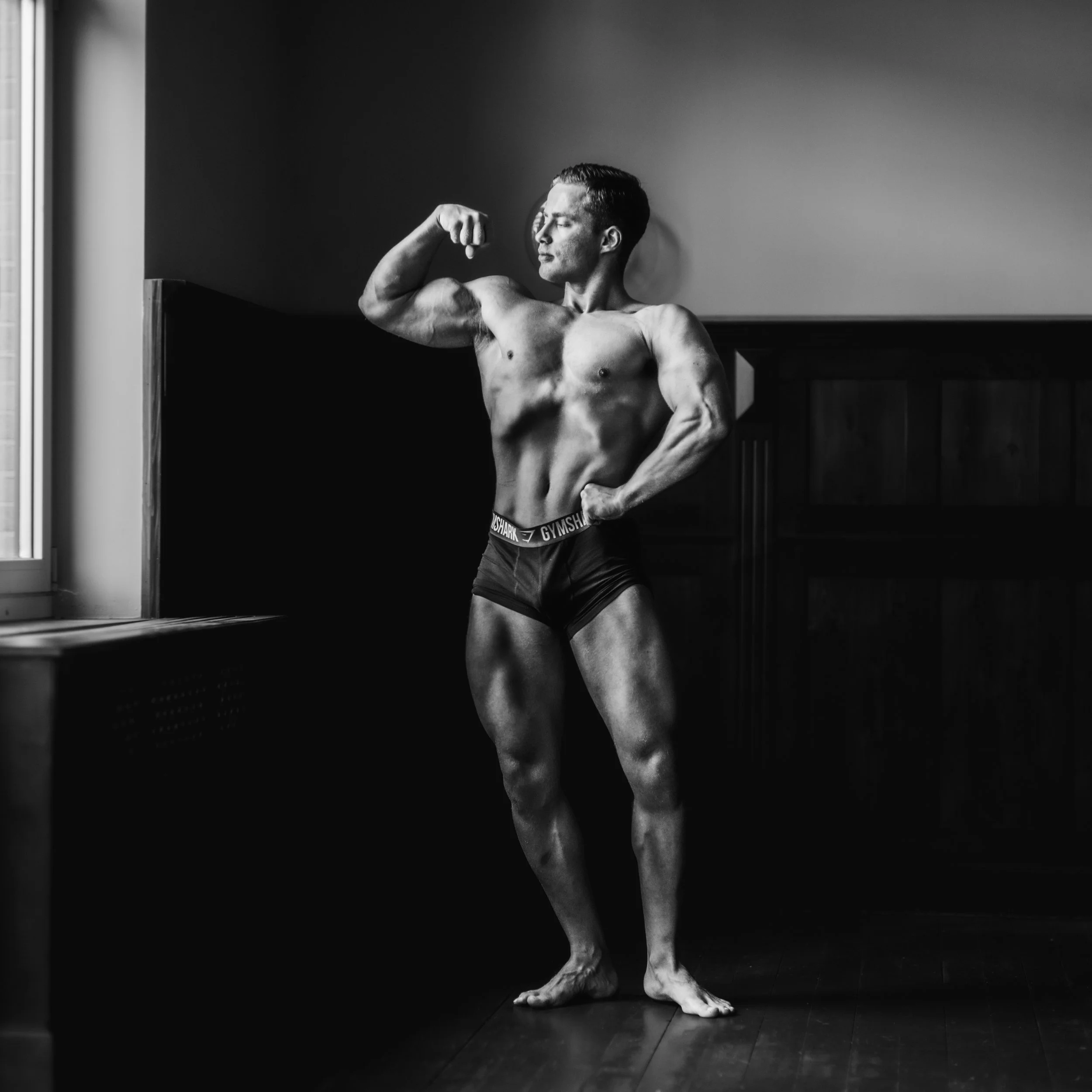 Black and white photo of a shirtless man with a lean, muscular physique posing with his left arm flexed and his eyes closed, standing against a plain wall near a window with vertical blinds.