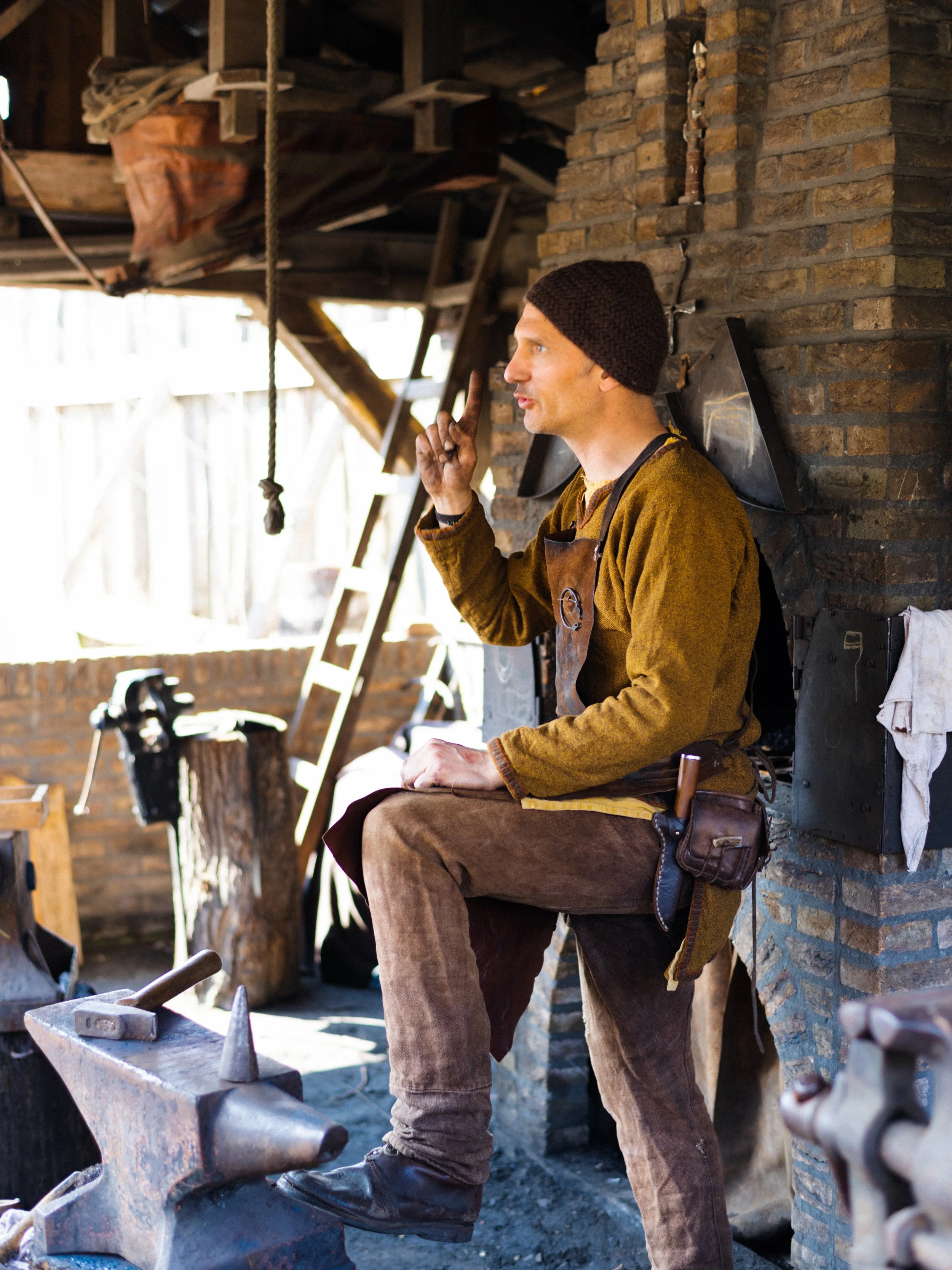 A blacksmith wearing a brown apron and a knit cap, sitting on a stool, with an anvil and hammer in front of him inside a rustic workshop with brick walls.
