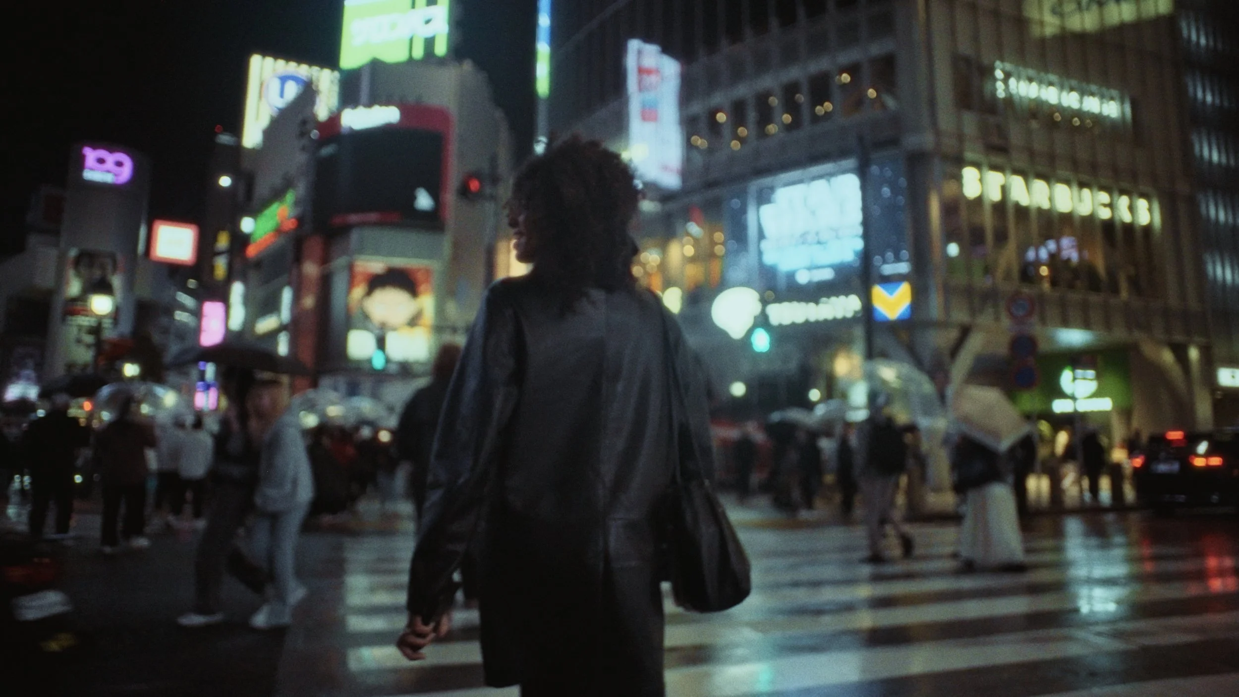 A person with curly hair wearing dark clothing standing at a busy crosswalk at night in an urban area with bright neon signs.