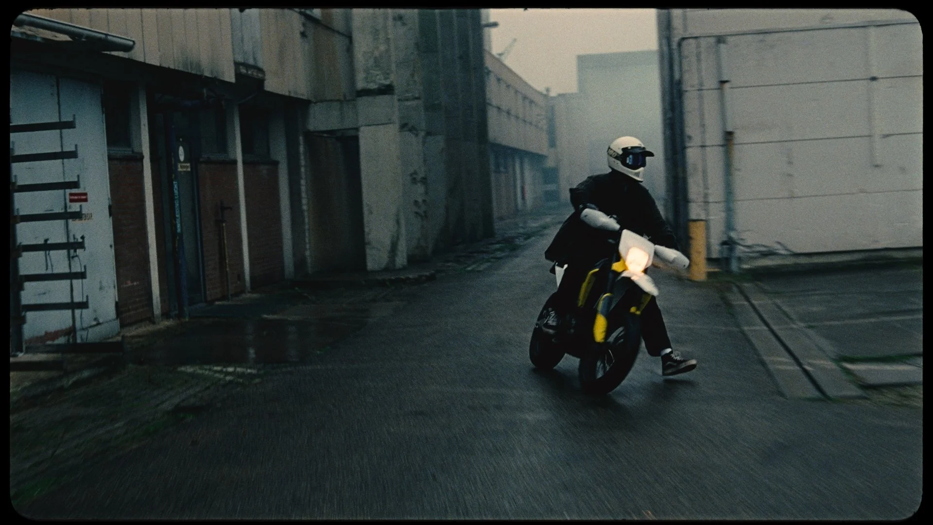 A person riding a small motorcycle or scooter down a narrow, deserted street in a foggy or rainy urban environment, wearing a helmet and dark clothing.