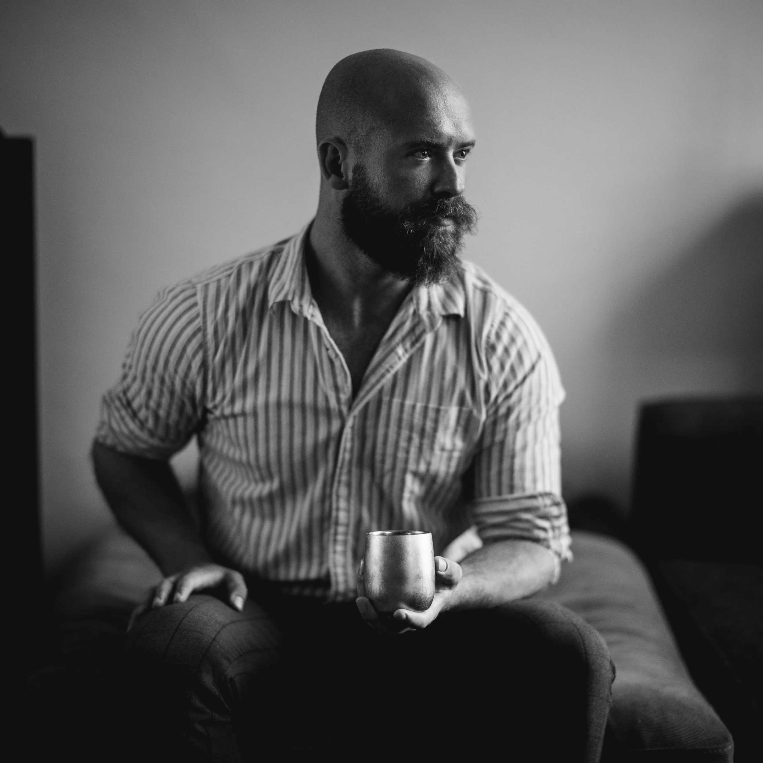 Black and white photo of a man with a beard and shaved head sitting cross-legged, holding a cup with both hands, wearing a striped shirt and jeans, in an indoor setting.
