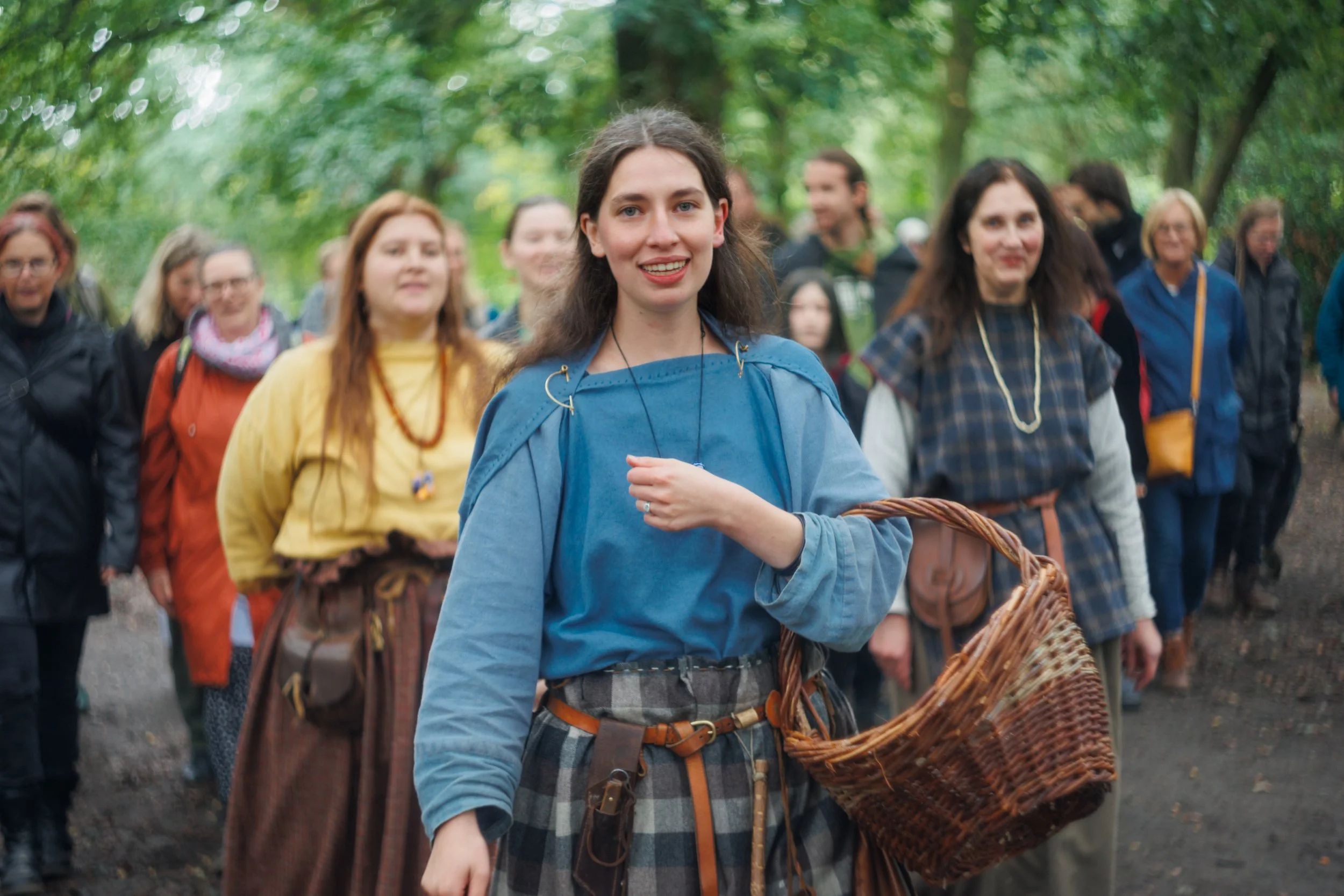 A young woman with dark hair and a blue jacket smiling and holding a woven basket while walking in a forest with a group of people.