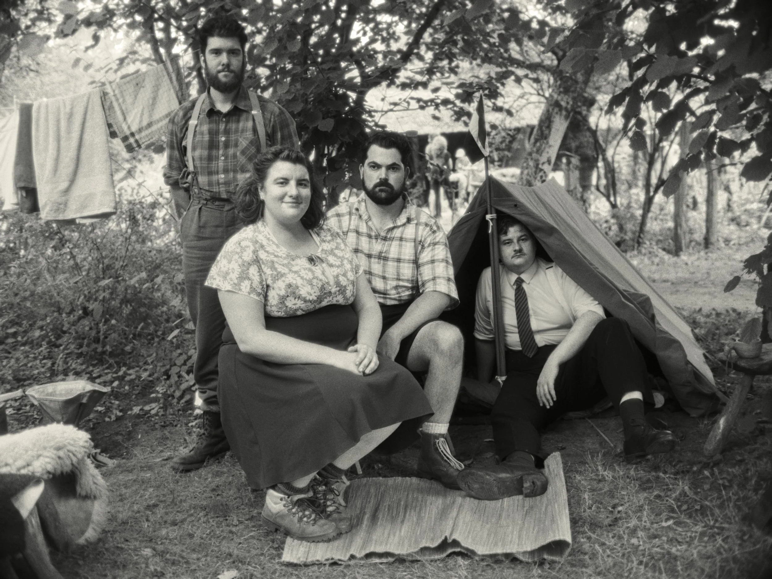 A black-and-white photo of five people outdoors in a wooded area with trees in the background. One woman with shoulder-length hair and a floral blouse is sitting on a log. To her right, a man with a beard, dark hair, and a checkered shirt is sitting 