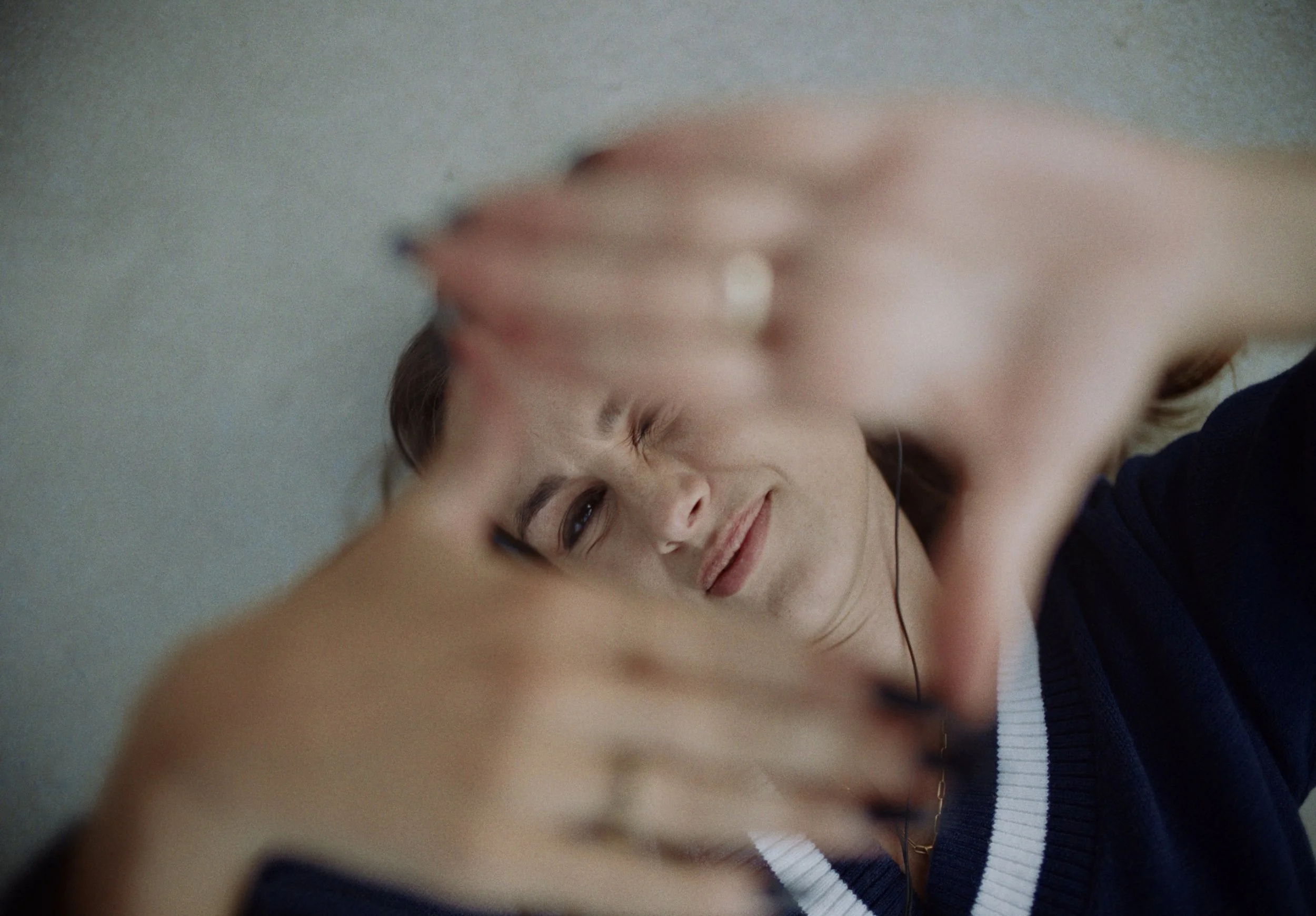Close-up of a smiling woman with brown hair and light skin, lying down with her arm raised above her head, wearing a navy blue and white striped top, against a plain background.