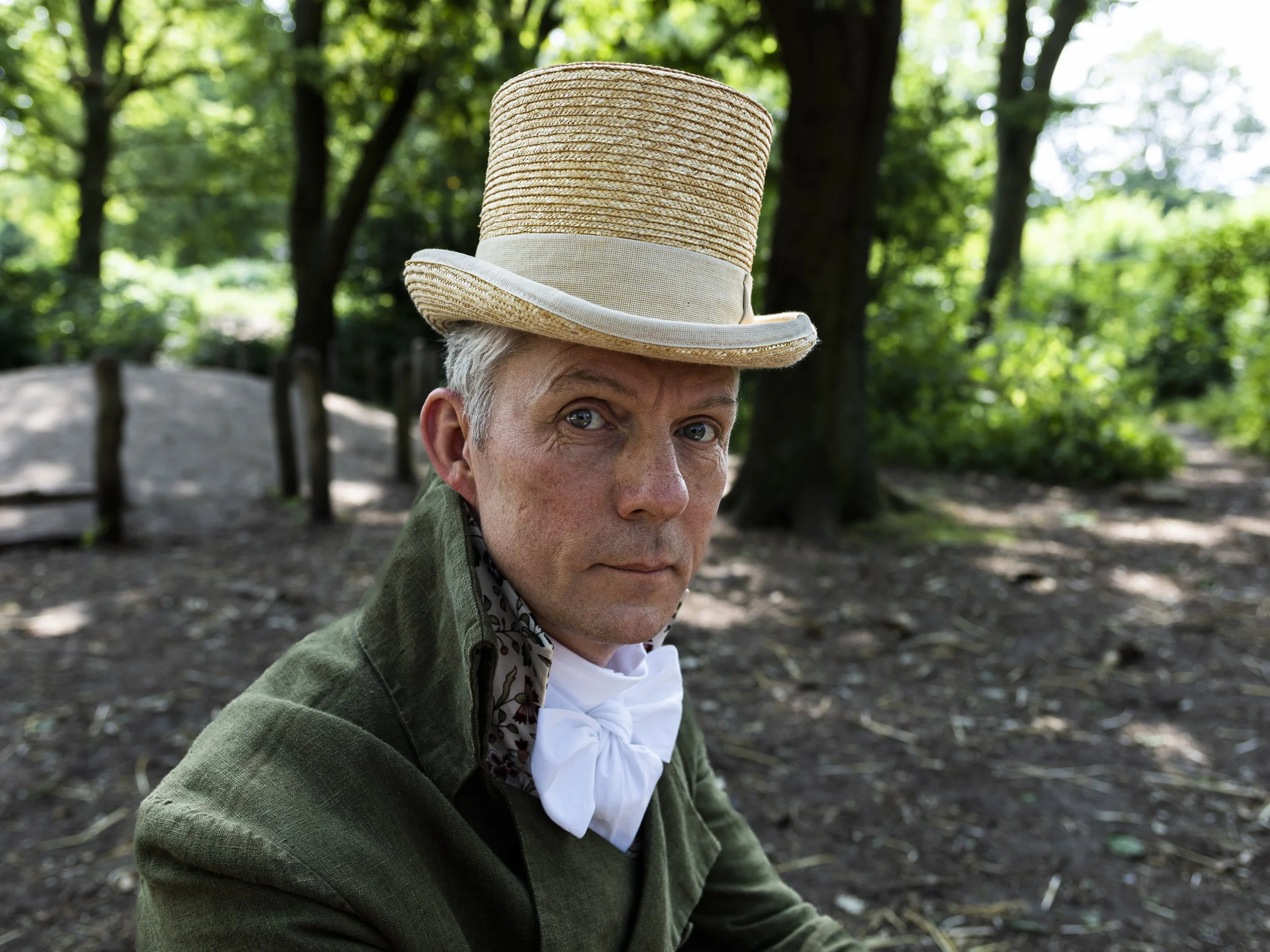 A man with gray hair and blue eyes wearing a large straw hat with a beige band, a green jacket, and a white shirt with a bow, sitting outdoors in a park with trees and sunlight filtering through the leaves.