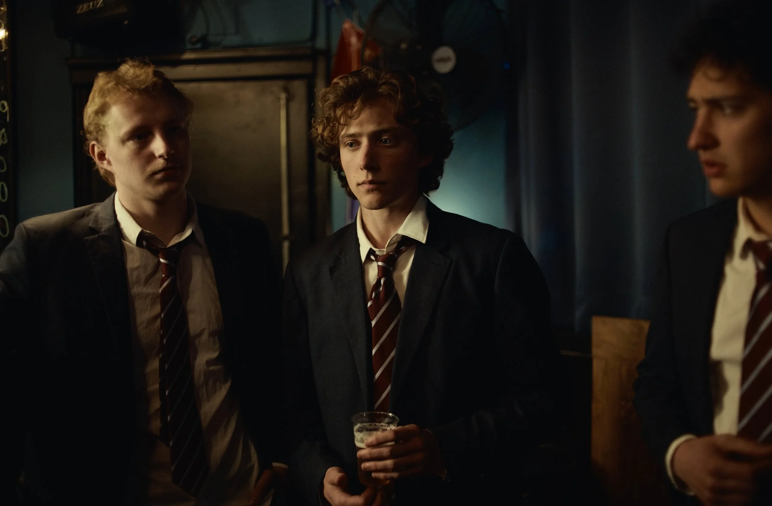 Three young men dressed in black suits, white shirts, and striped ties standing indoors, with one holding a glass of beer.