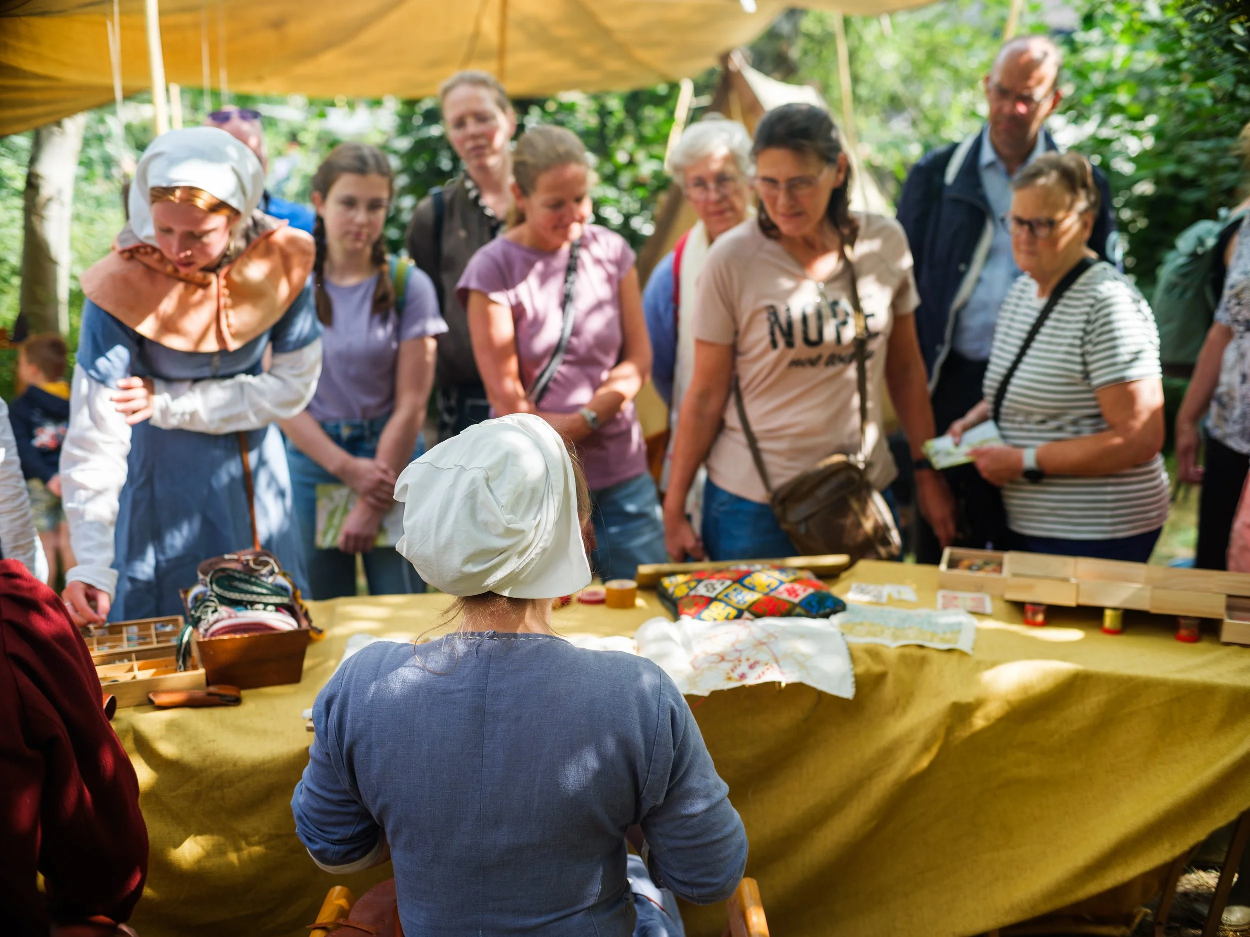 Group of people gathered around a table outdoors, looking at items on display at a market or fair under a tent with trees in the background.