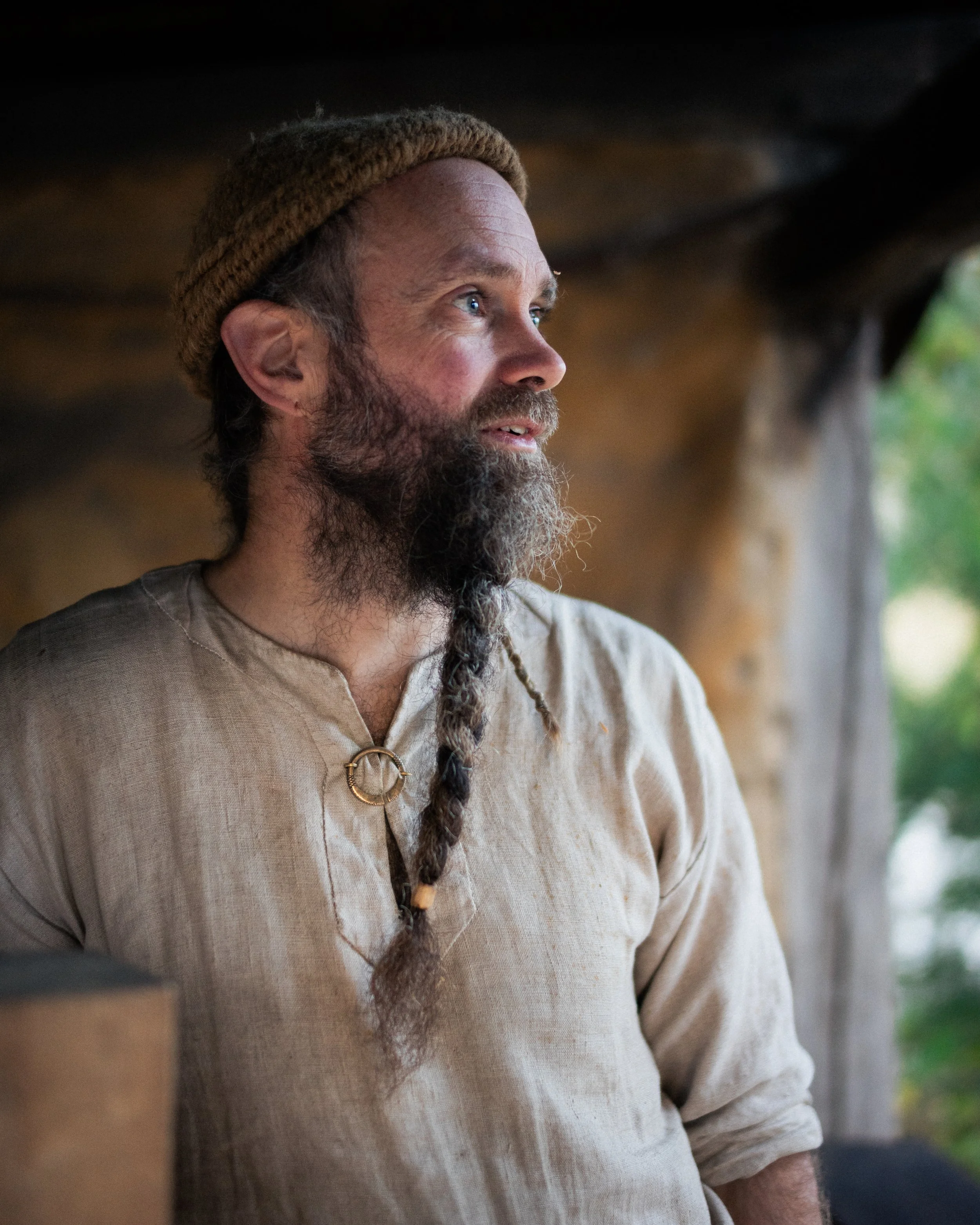 A man with a beard wearing a brown knit hat and beige linen shirt, looking thoughtfully to the right, with blurred background of wooden structure and green foliage.