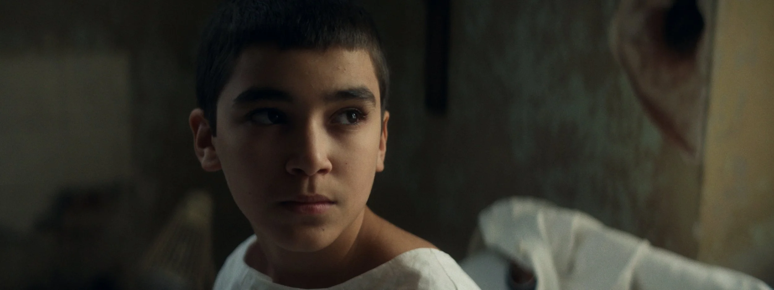 Close-up of a young boy with short dark hair, looking thoughtfully into a mirror in a dimly lit room.