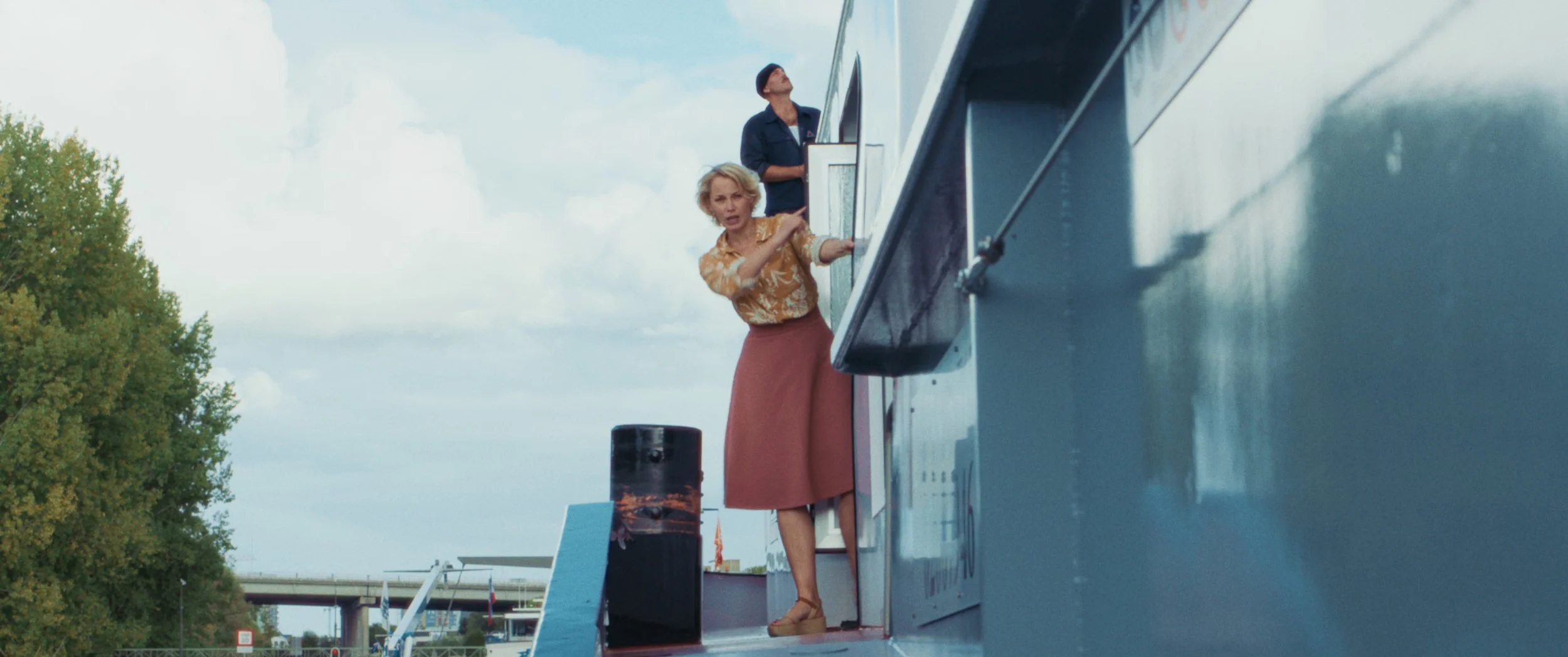 A woman and a man on a blue boat, with the woman standing at a window and the man looking out, during daytime with a partly cloudy sky.