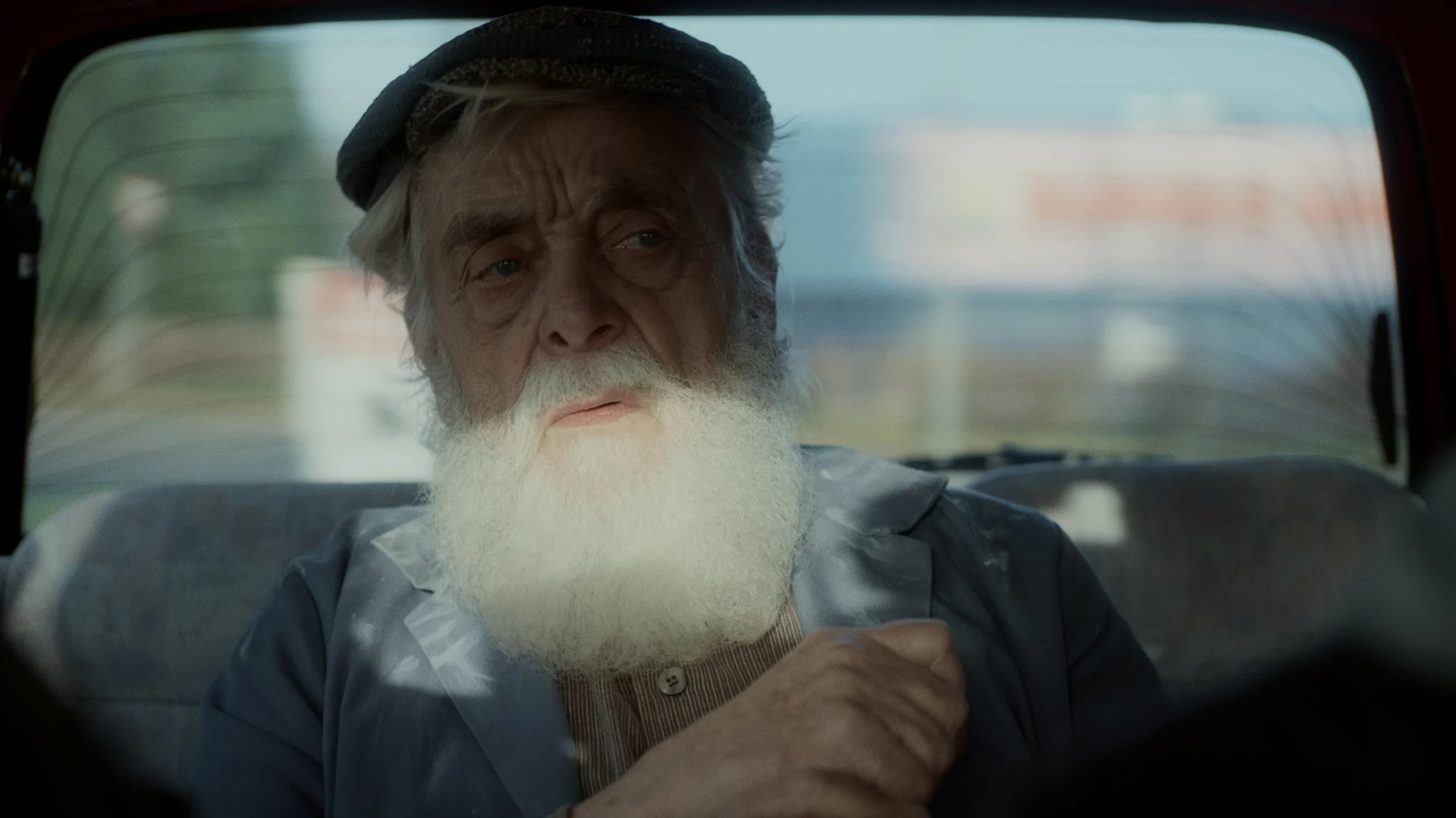 An elderly man with a white beard and mustache wearing a gray hat and gray jacket is sitting in the backseat of a vehicle, looking out the window.